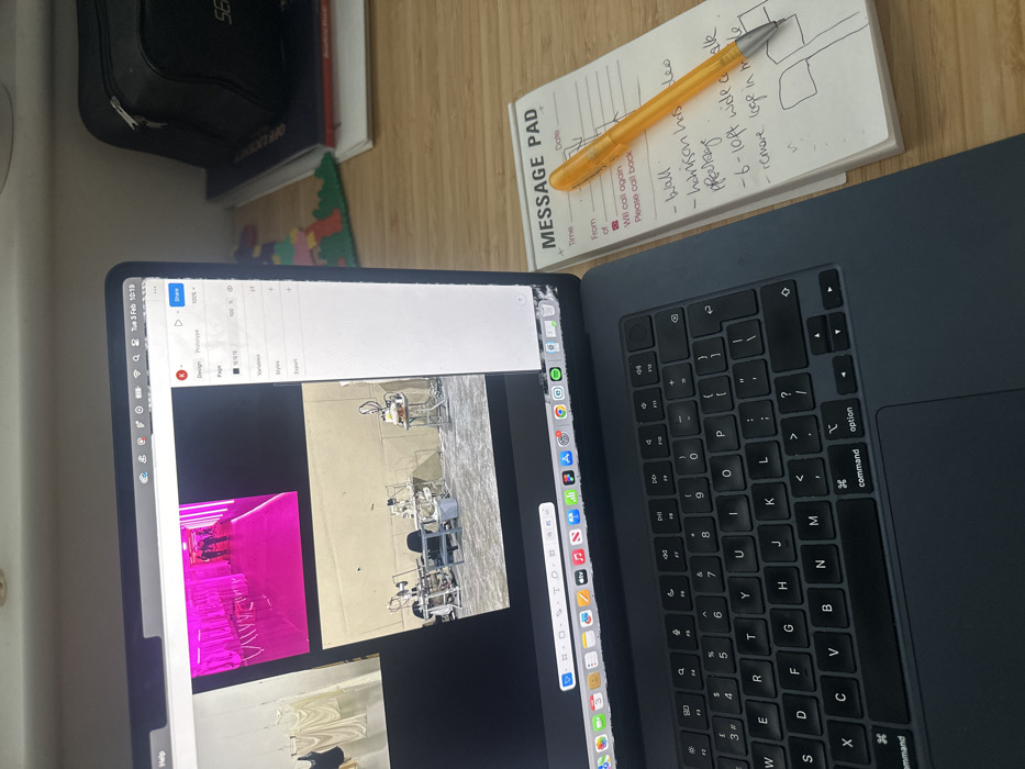 The image depicts an indoor workspace, likely an office or home office environment, positioned on a light-colored wooden desk. The specific location in Wealden, United Kingdom, cannot be determined from the visual information provided.
The primary subject is a dark grey Apple MacBook laptop, featuring a black keyboard and trackpad. The laptop screen displays a graphical user interface for design software, with "Design" and "Prototype" tabs visible in the right-hand panel. The top menu bar shows "Tue 3 Feb 10:19". The screen exhibits minor physical damage in the bottom right corner. Within the software's canvas, three distinct images are visible:
1. A top image showing a corridor or room bathed in bright pink light, with two indistinct human figures in the distance.
2. A large central image depicting an industrial or workshop-like setting with a concrete floor and plain walls. Multiple pieces of machinery or equipment are arranged on stands and a table, alongside a blue bucket.
3. A partial image on the far left showing white, draped fabric items hanging, possibly clothing or textiles.
To the right of the laptop is a white "MESSAGE PAD" notepad with an orange transparent ballpoint pen resting on it. Handwritten notes are visible on the pad, including bullet points such as "- wall", "- harrison hots video", "- ppdge", "- 6-10ft wide aisle", and "- remove loop in middle," along with a simple drawing resembling two intersecting axes or poles at the bottom.
In the background, on the desk surface behind the laptop, a black fabric bag, several stacked books, and a colorful plastic building block structure (possibly LEGO) are visible. No people are present in the foreground of the scene.