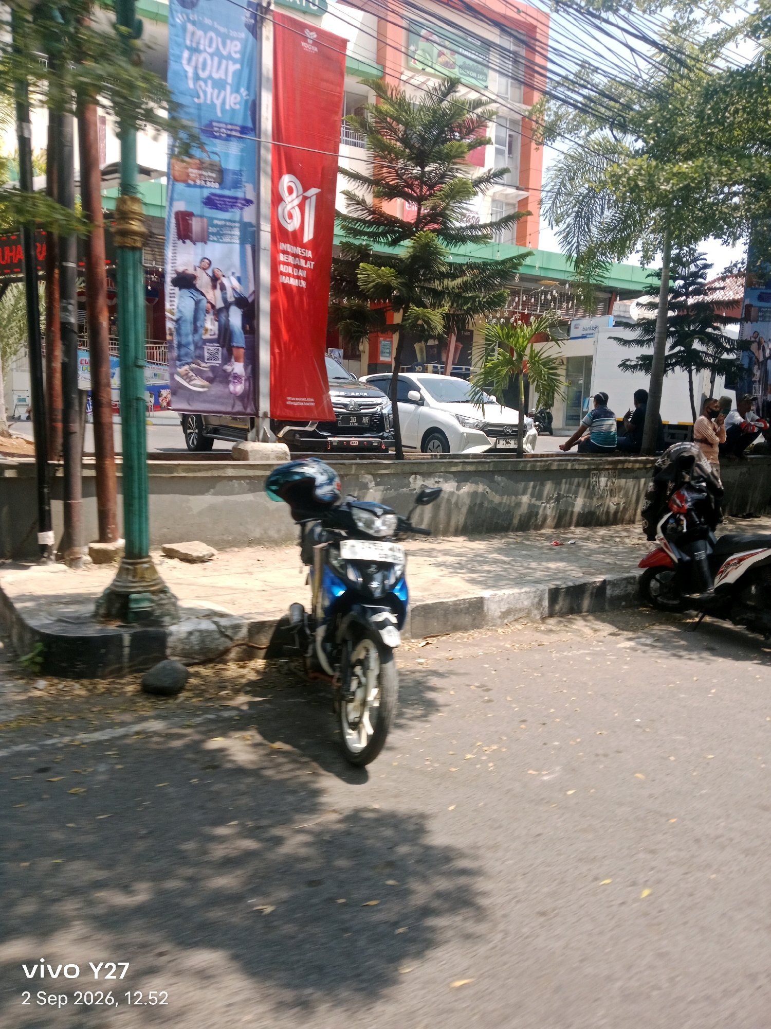 The image shows a street scene in Palabuhanratu, Indonesia, with two large banners hanging vertically. One banner is blue and features the text "move your style" and images of luggage and people. The other banner is red and displays the number "81" in large white font, along with text in Indonesian. In the foreground, there are two motorcycles parked on the side of the road. The street is paved and there are trees and buildings visible in the background. Several people are seated on a low wall on the sidewalk, some appearing to be resting or observing the surroundings. The overall impression is that of a typical urban street in a tropical climate.