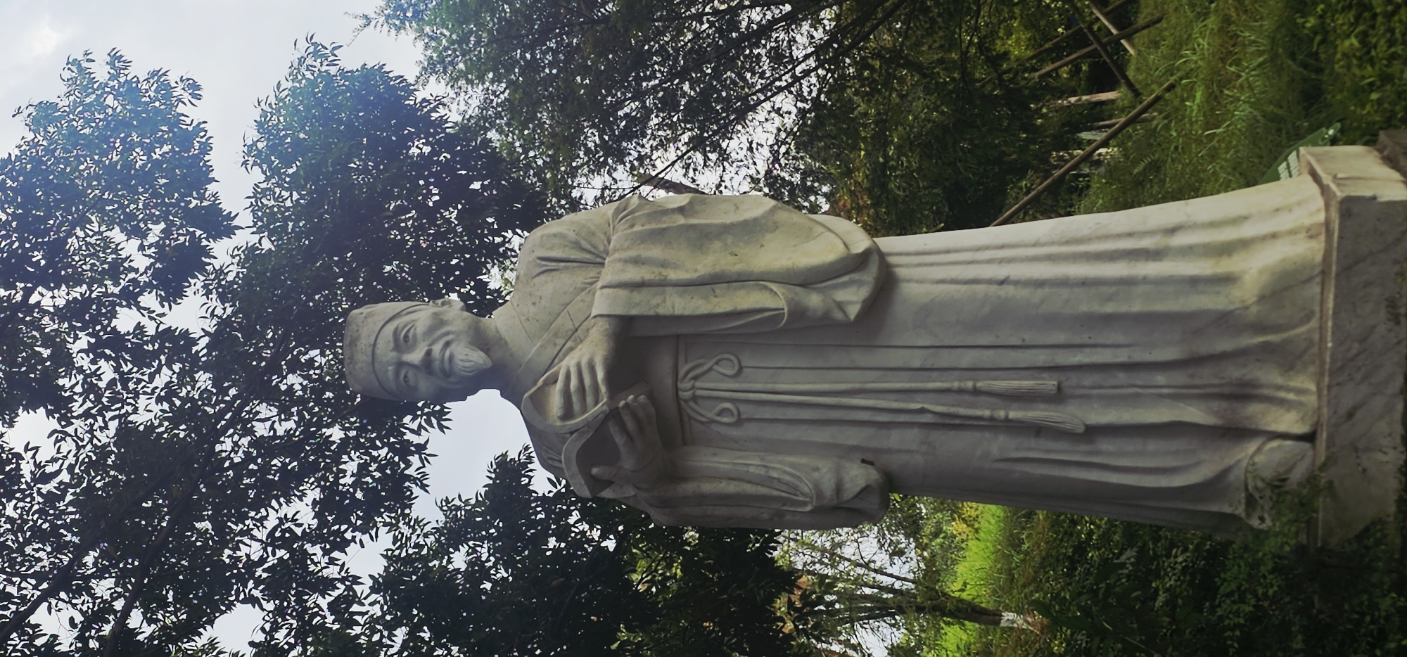 A full-body, low-angle shot captures a weathered, light-grey stone statue of an elderly East Asian man outdoors. The statue depicts the man wearing traditional robes and a cap, with a long beard and mustache. He holds an open book in his hands, which are clasped together. The statue stands on a stone pedestal. Lush green foliage and trees form the background, with sunlight filtering through the leaves. The setting appears to be a park or garden area. The location is identified as the Chongqing High-tech Zone, China.