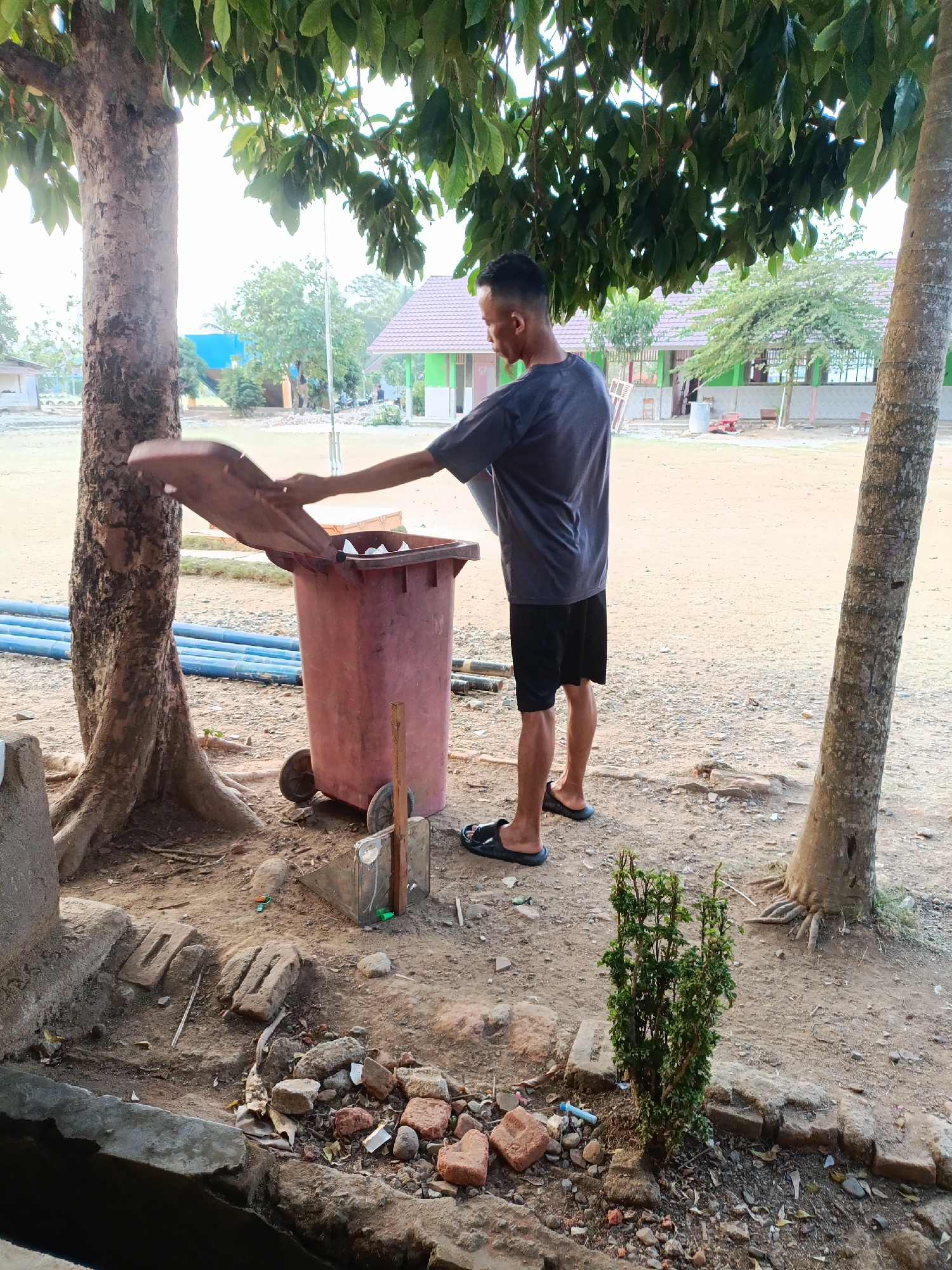 In Sumberejo, Indonesia, a young man stands near a large, weathered, reddish-brown trash bin under the shade of a tree. He is wearing a dark grey t-shirt and black shorts, with black flip-flops on his feet. The man is holding the lid of the trash bin open with his right hand, appearing to be in the process of discarding something into it. The trash bin has two wheels and looks somewhat worn. Behind the man and the trash bin, there is a sandy, uneven ground with scattered debris, including rocks and broken bricks. Further in the background, there are buildings with green roofs and some trees. Several long blue pipes are visible on the ground to the left of the trash bin, partially obscured by a large tree trunk. A small, green leafy bush is planted to the right of the trash bin, near another tree. The overall setting appears to be an outdoor area, possibly a schoolyard or a public space.