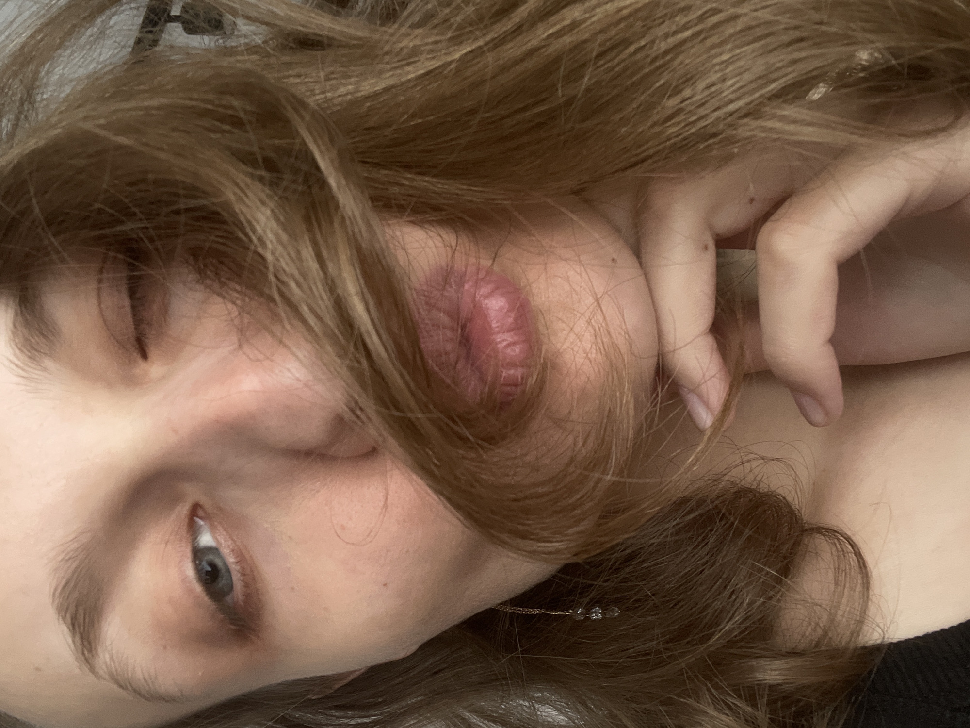 A close-up, angled headshot shows a person with fair skin and light brown, wavy hair. The person's right eye is partially open, revealing a grey iris. A hand with light skin is positioned near the person's face, fingers slightly curled. The lips are prominent and appear full. An earring with small clear stones is visible near the ear. The location, Подшивалово, Russia, is not discernible from the image.