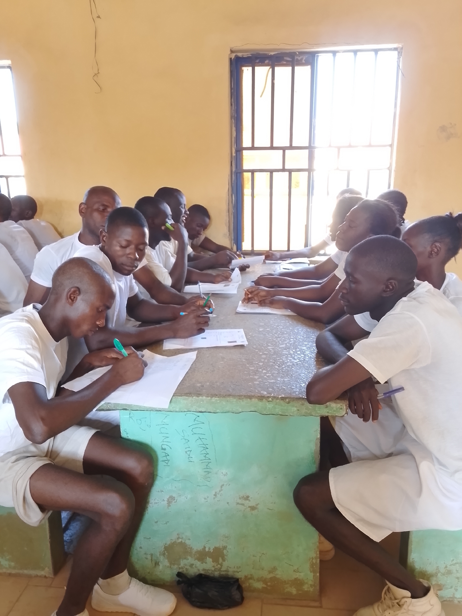 The image depicts an indoor scene resembling a classroom or examination hall, likely situated in Kaduna, Nigeria. Approximately a dozen individuals, predominantly young adult males and some females, are seated at two long, worn light-green tables. They are all uniformly dressed in white short-sleeved shirts and white shorts or light-colored trousers, suggesting an institutional setting such as a school, training facility, or possibly a correctional institution.
The individuals are actively engaged in writing or filling out forms, with papers and pens laid out before them on the tables. Their focused expressions indicate concentration, suggesting they are undergoing an examination, test, or an application process. Bright natural light streams into the room through large, metal-barred windows in the background, illuminating the scene and indicating it is daytime. The walls are a plain light yellow or beige.
On the side of the table in the foreground, faint graffiti is visible, including the names "MUHAMMAD," "MUNGADI," and "SA'IDU." The overall atmosphere is one of quiet diligence and structured activity within a utilitarian environment.