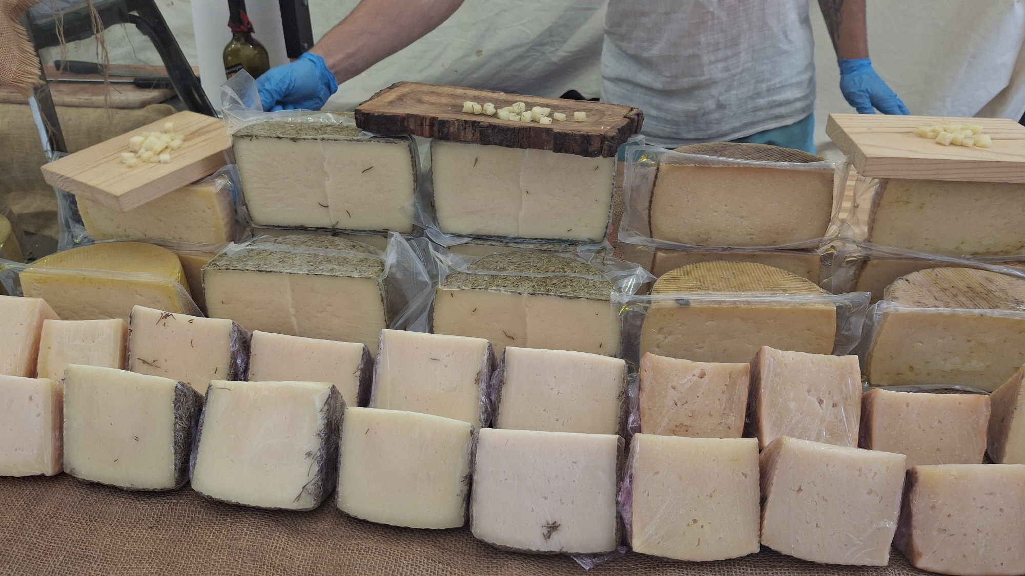 This image shows a display of various cheeses, likely at an outdoor market in La Fresneda / la Freixneda, Spain. In the background, a person wearing blue gloves is visible, interacting with the cheese display. The foreground is dominated by stacks of cheese wedges, some whole and others cut. The cheeses vary in shape, size, and rind, with some appearing to be aged and others fresher. They are individually wrapped in plastic. The cheeses are arranged on a table covered with a burlap-like material. A small wooden board with diced cheese pieces is placed on top of one of the larger cheese blocks. Behind the cheese, a partial view of a bottle of wine and some market stall elements are visible. The lighting suggests it is daytime.