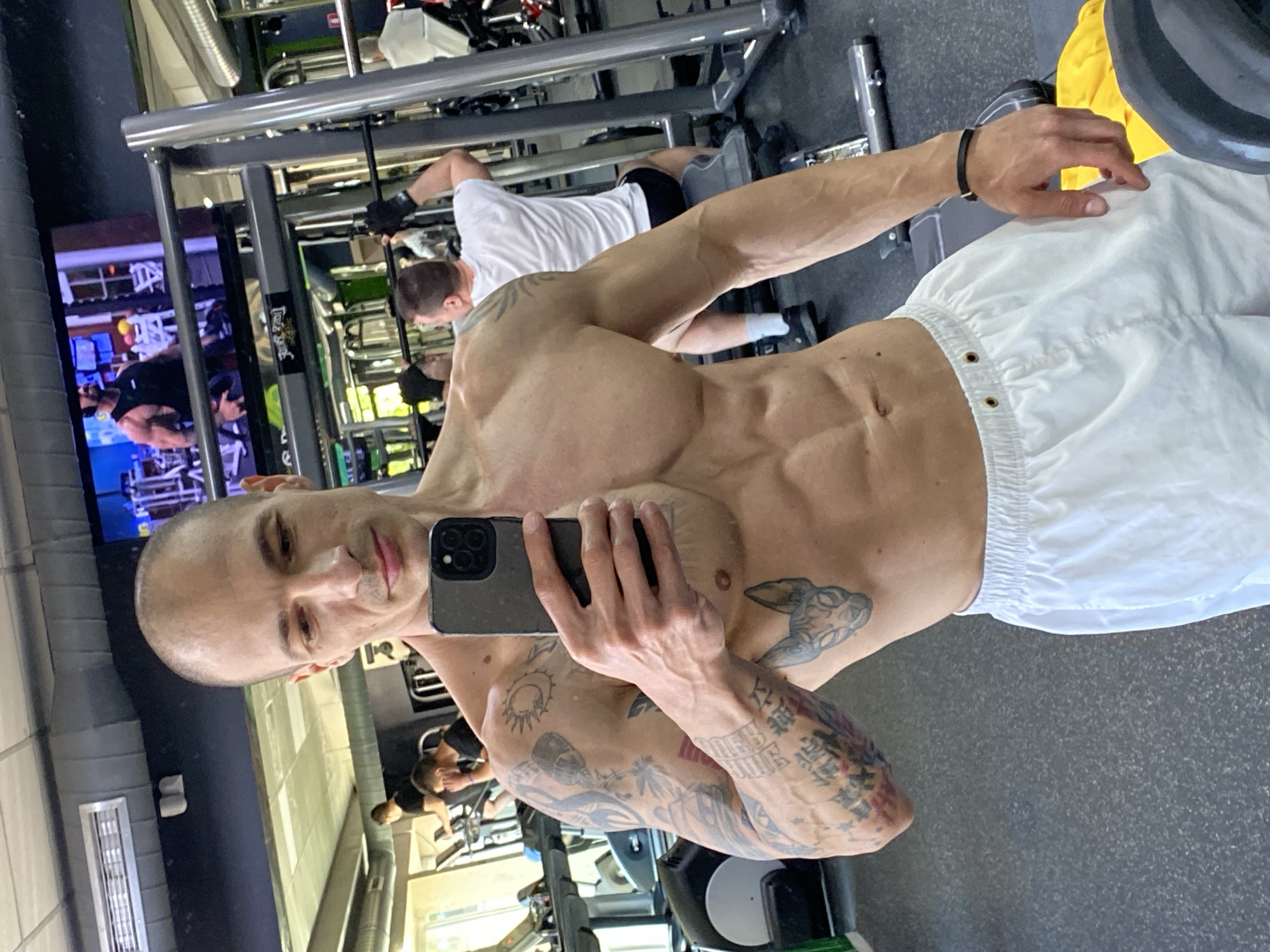 A shirtless man takes a selfie in a gym. He is bald and has tattoos on his arms, chest, and torso. He wears white shorts. The background shows other gym-goers and equipment. A television screen is visible in the background. The location is in a gym; there is no specific information that it is in Pleven, Bulgaria.
