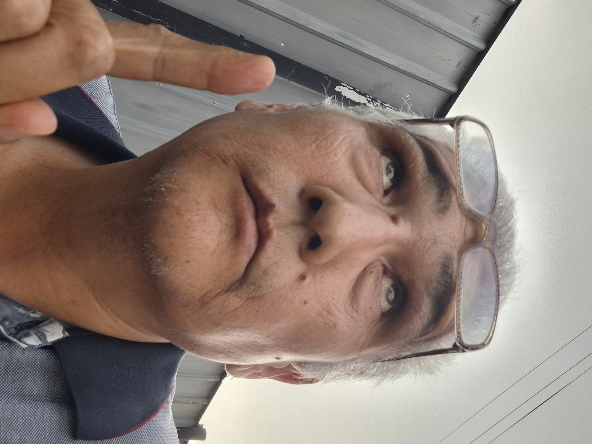 The image is a close-up, low-angle selfie of a man looking upwards. He is wearing glasses perched on his forehead, revealing his eyes which are looking up and to the left. His mouth is slightly open, and his chin has some stubble. His right hand is raised, with his index finger pointing upwards. He is wearing a collared shirt, partially visible. The background shows a corrugated metal structure on the left and a cloudy sky with power lines in the distance. No specific location in Almaty, Kazakhstan is discernible from the image.