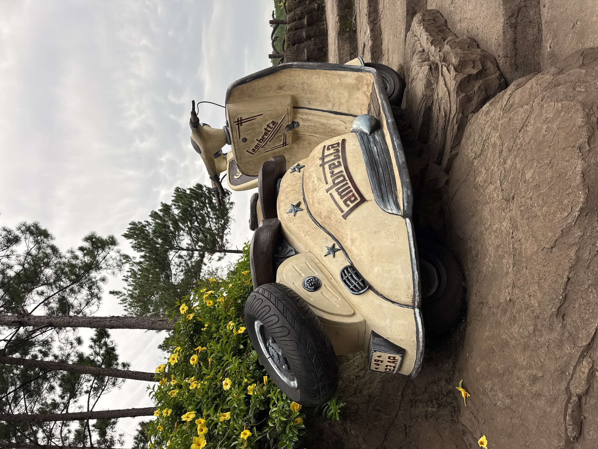 This is a full, slightly low-angle, eye-level shot of a vintage cream-colored Lambretta scooter sculpture. The scooter is positioned on a series of tiered, rough, brown stone steps, with a backdrop of lush green bushes with bright yellow flowers and tall pine trees under a cloudy sky. The scooter is the primary subject, taking up most of the frame. It has a spare tire mounted on its rear, and the name "Lambretta" is prominently displayed in a stylized font on both the rear fender and the front leg shield. The scooter has some decorative stars on its rear. A license plate on the rear reads "49 123 AP." The stone steps appear worn and weathered. The environment suggests an outdoor setting, possibly a garden or park. The scene is static; there are no people or animals present. The lighting is diffused due to the overcast sky, creating soft shadows. The overall impression is one of a rustic and nostalgic display, potentially in a tourist location. This appears to be a Lambretta sculpture located at the Dinh Cau lighthouse viewpoint in Da Lat, Vietnam, where it is a popular photo opportunity.