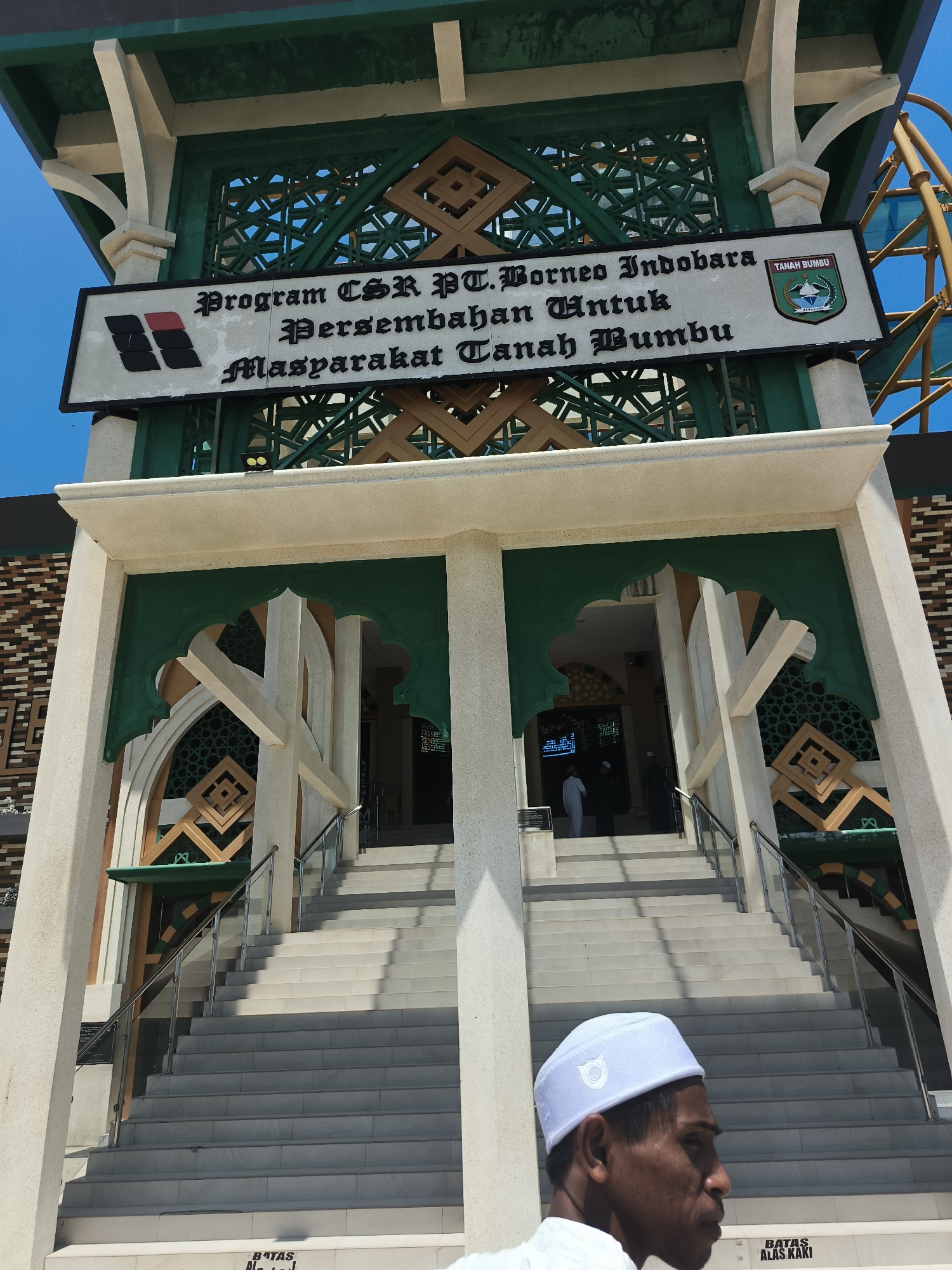 A man wearing a white peci cap stands in the foreground, facing right, looking towards a building. Behind him, a wide set of concrete stairs leads up to the entrance of a building adorned with green and beige architectural elements, featuring Islamic geometric patterns. A sign above the entrance reads "Program CSR PT. Borneo Indobara Persembahan Untuk Masyarakat Tanah Bumbu," with the logo of Tanah Bumbu regency on the right. The sky is clear and blue. The overall scene appears to be in a public area, possibly a mosque or a community building, located in Tanah Bumbu Regency, Indonesia.