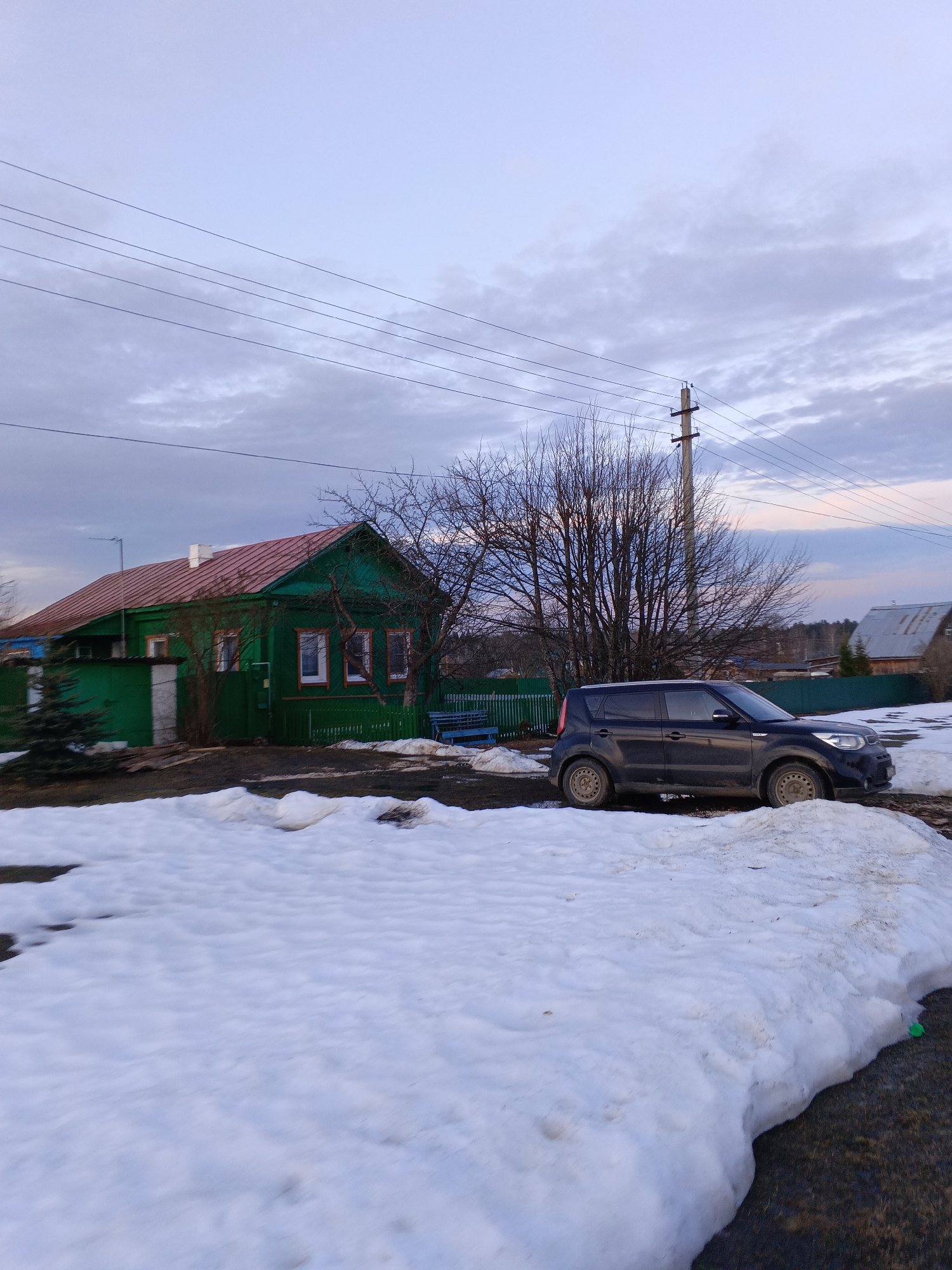 The scene depicts a cloudy early spring or late winter day in Красный Маяк, Russia. A dark blue Kia Soul is parked on a patch of mud and snow. In the background is a traditional green Russian wooden house with a red roof. A utility pole with wires is visible behind the vehicle and leafless trees. The ground is covered in melting snow.