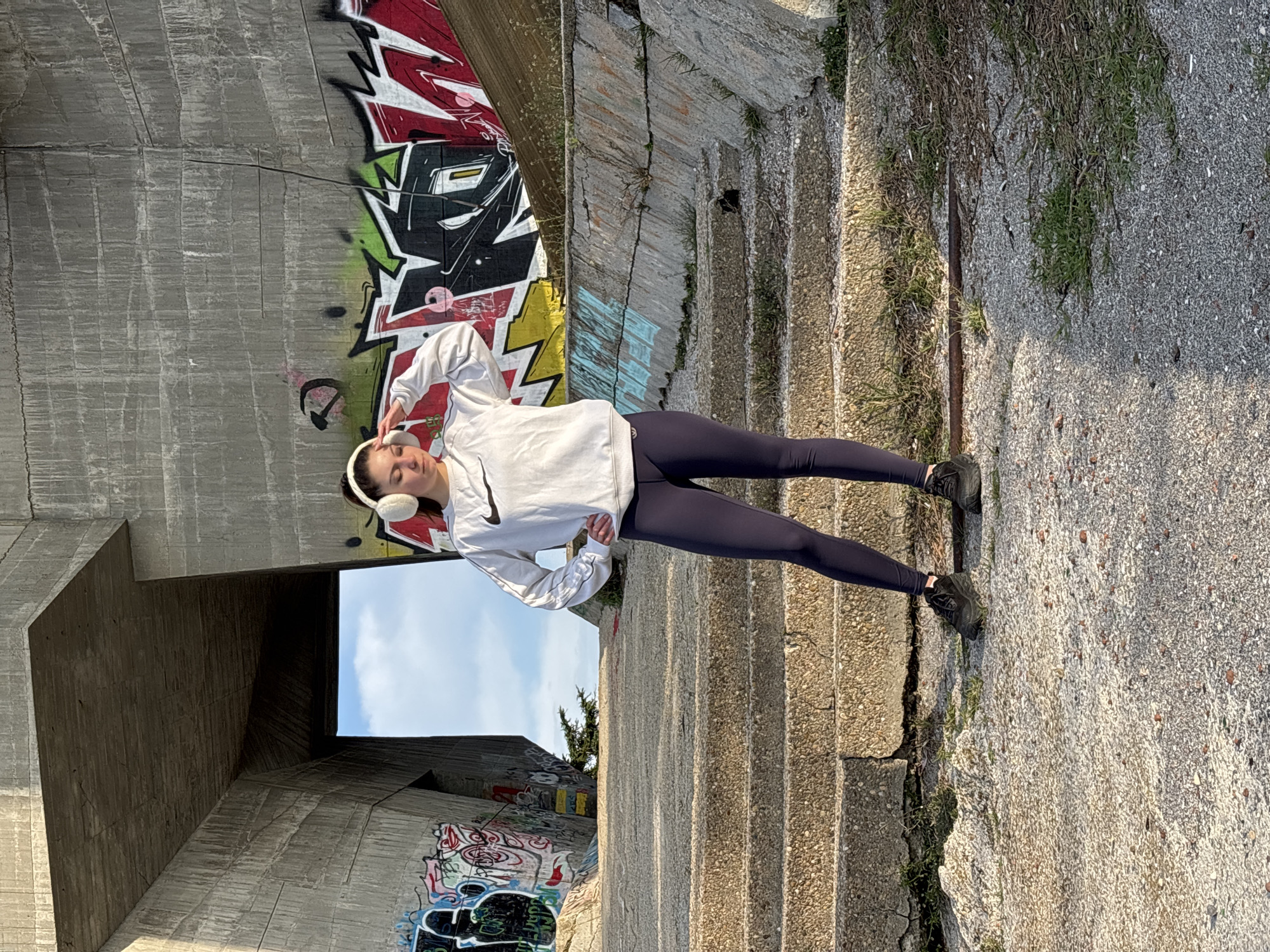 A person in athletic wear and headphones stands on the side of a set of concrete stairs with graffiti on the adjacent walls. The person is wearing a white Nike sweatshirt and dark leggings. They are facing forward with their legs straight and their arms bent at the elbows. The stairs are made of rough concrete and some have grass growing in the cracks. The graffiti on the walls includes a variety of colors and designs. There is a blue sky visible through an opening in the structure.