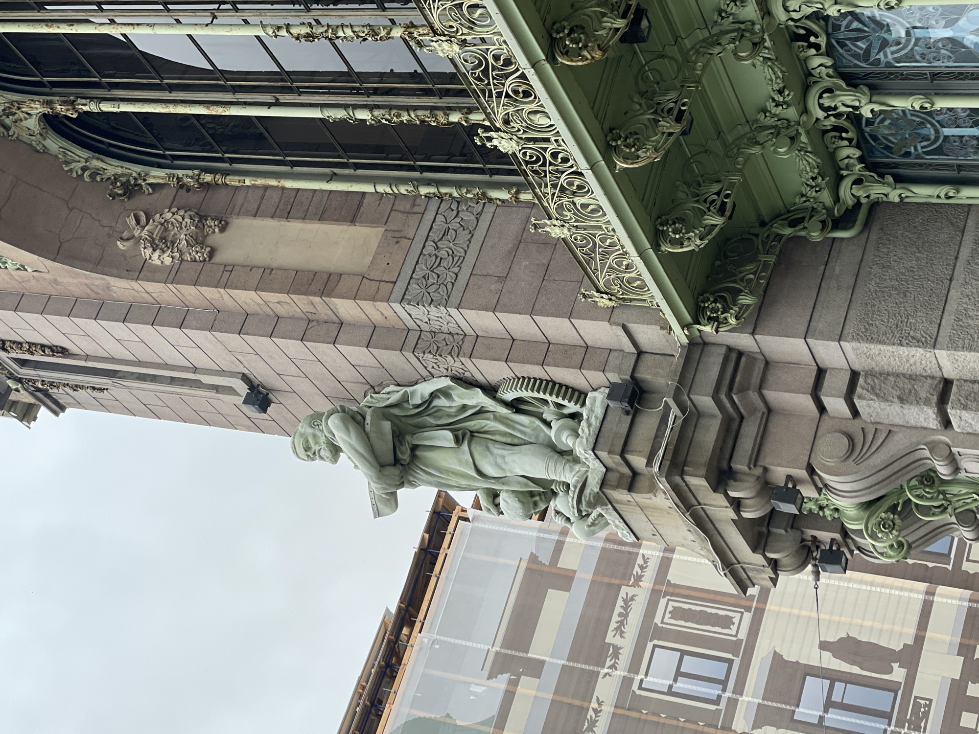 The image shows a portion of the facade of a building in Saint Petersburg, Russia, specifically part of the Singer House (also known as the Dom Knigi or House of Books) located on Nevsky Prospekt. The view is taken from a low angle, looking upwards at the building. The architectural details include a light-colored stone facade, with decorative elements such as floral patterns, and a statue of a male figure with books and a gear wheel. Above, there is a section with green metalwork and glass. The lower part of the frame includes a section of the building’s facade which is covered with scaffolding and a scrim that features a print of the building's own architectural details. Wires are also visible, running along the facade. The sky is overcast.