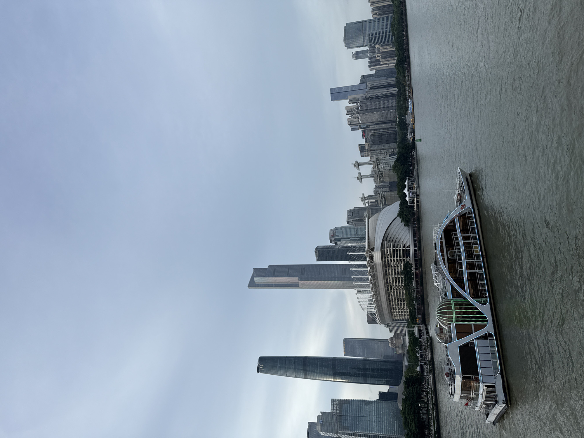 The image captures a view of the Haizhu District in China, featuring a modern cityscape and a prominent river. In the foreground, a large, multi-decked tour boat with illuminated accents moves across the murky water. The boat is sleek and contemporary, with large windows and decorative lighting.
Beyond the river, the Haizhu District skyline is dominated by a collection of modern skyscrapers of varying heights and architectural styles. Notably, two particularly tall and distinctive towers are visible: one with a rounded, tapering form and the other a sharp, angular structure. Between these towers and extending outwards, a dense cluster of office buildings and residential towers forms the urban landscape. In the mid-ground, a large, white, curved structure with a visible support system, possibly a stadium or convention center, sits adjacent to the riverbank. The sky above is overcast, with a pale, muted light casting a soft glow over the scene. Trees line the riverbank in some areas, adding a touch of greenery to the urban environment.