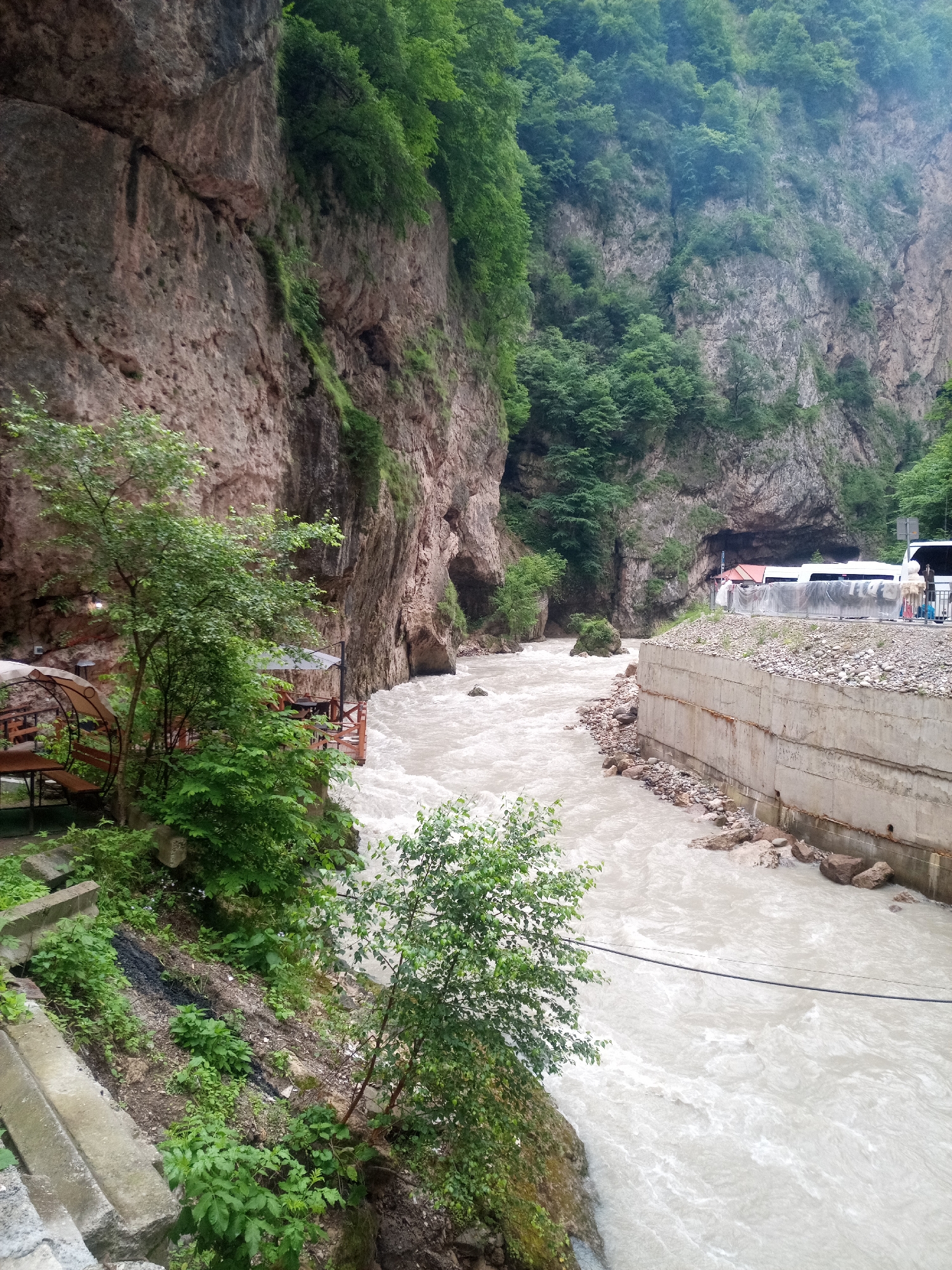 The image depicts a wide, fast-flowing river carving through a narrow gorge in Khushto-Syrt, Russia. The river's water is milky-white, indicating a high sediment load, and churns with visible rapids. Sheer, steep rock faces covered in lush green vegetation rise on both sides of the gorge. On the left bank, a rustic wooden deck with tables and seating is visible, suggesting a potential rest stop or eatery. A few trees and dense green foliage grow along the riverbank and up the rocky slopes. On the right bank, a concrete retaining wall runs along the river, topped with gravel and rocks. Beyond the wall, a paved area is visible with two white buses parked side-by-side and several people standing near them. The sky is overcast, contributing to the muted lighting of the scene.