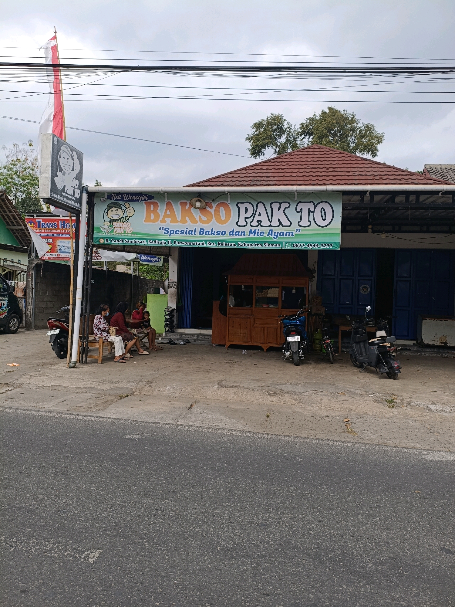 The image shows a storefront in Purwomartani, Indonesia, that appears to be a food establishment named "BAKSO PAK TO". The storefront features a prominent sign with the establishment's name and slogan, "Spesial Bakso dan Mie Ayam". To the left of the main sign is a smaller sign with a cartoon character and the words "Asti Wonogiri". There are three people sitting on a wooden bench outside the establishment, two women and a child. A motorcycle is parked to the left of the bench, and two other scooters are parked in front of the storefront. A wooden food stall is visible to the left of the entrance. The environment is an outdoor setting with a cloudy sky. The foreground is a paved road.
