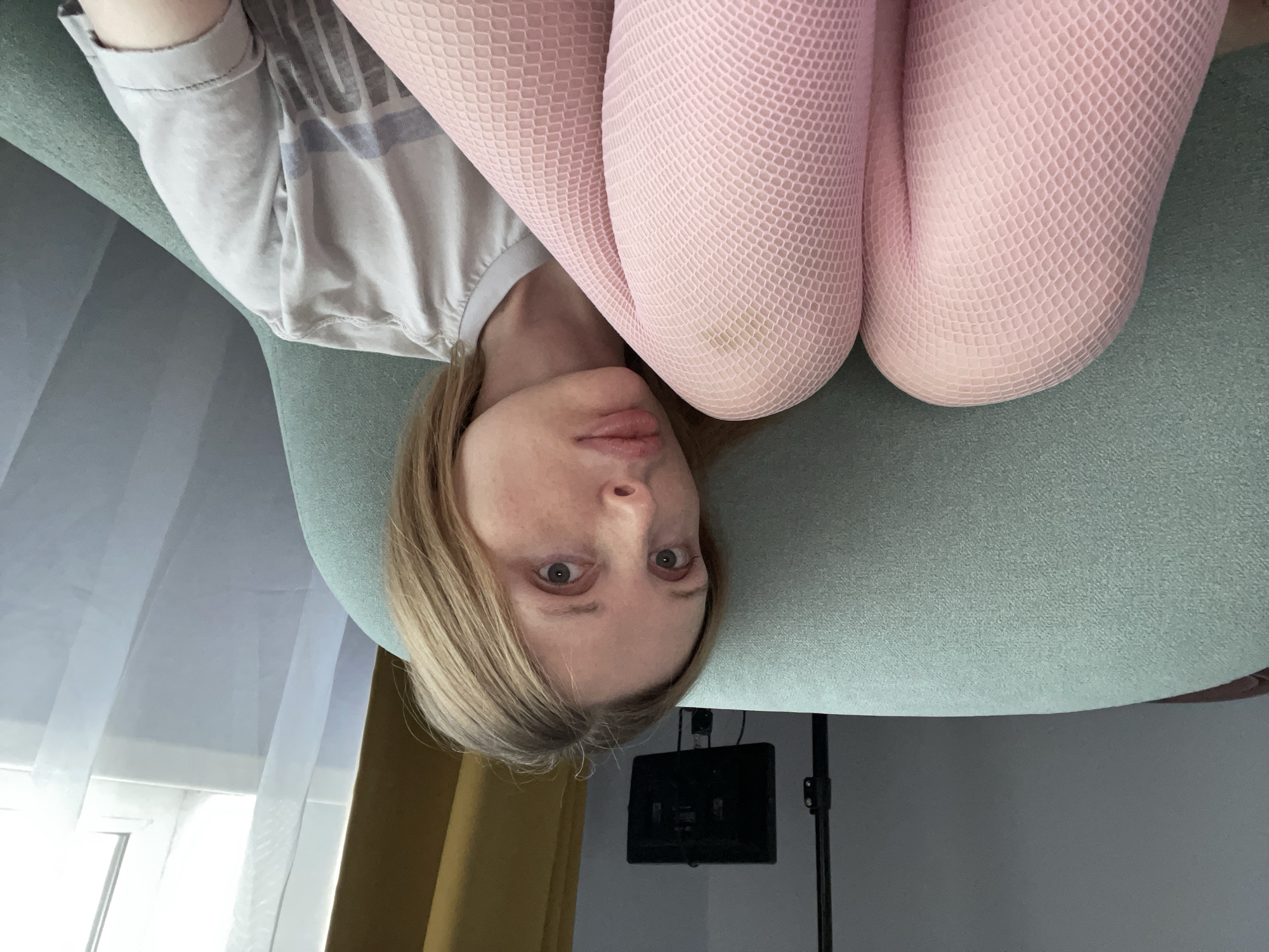 A woman with blonde hair and fair skin is sitting on a teal armchair, facing forward. She is wearing a light-colored t-shirt and pink fishnet tights. Her legs are crossed, and her knees are visible. The background includes a gray wall, a yellow curtain, and a window with sheer white curtains. A black lighting stand and a small black monitor are visible on the wall behind her. The scene appears to be an indoor setting, possibly a living room or studio, in Yablonovskiy, Russia.