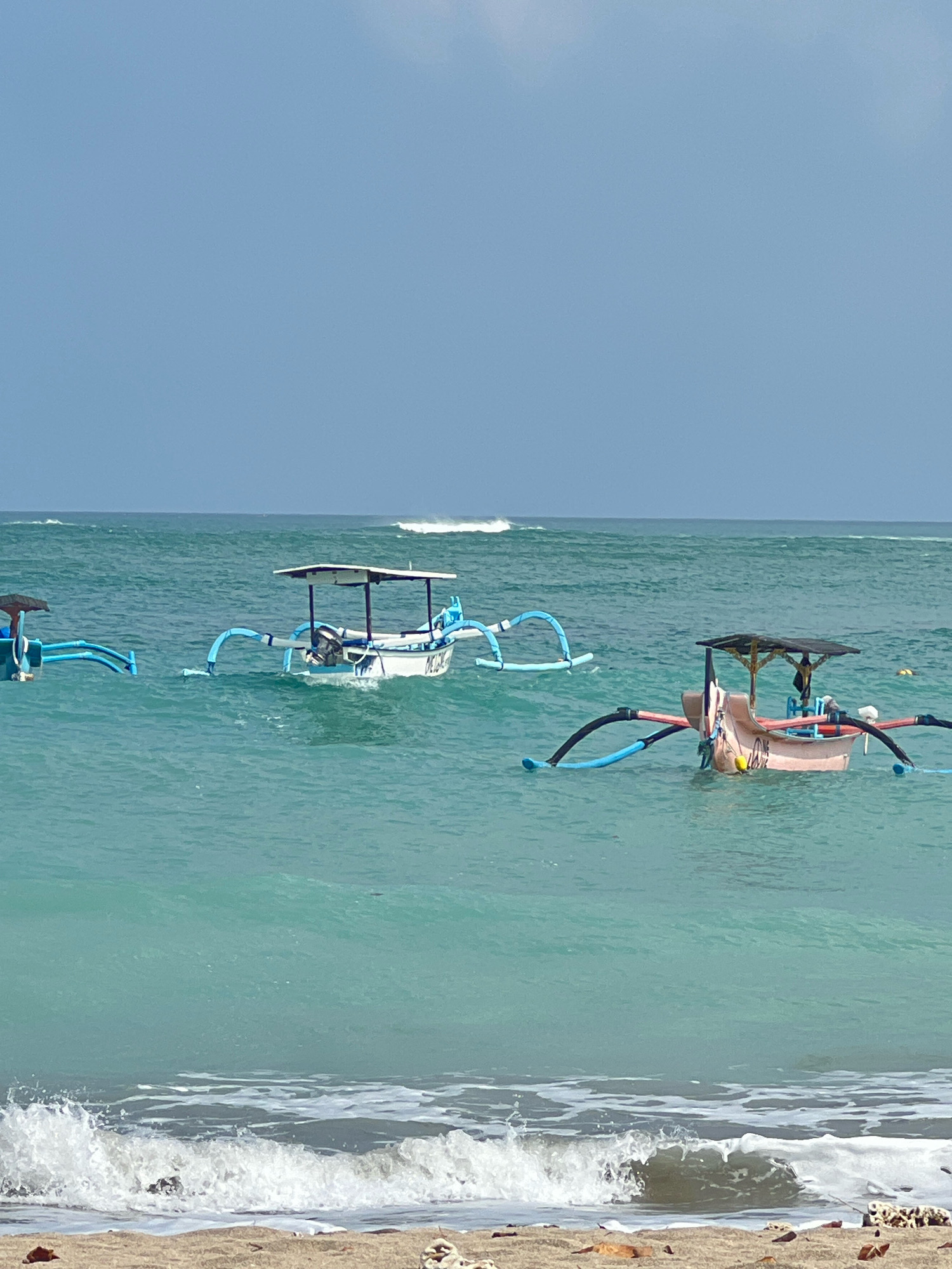 Three traditional Indonesian fishing boats, known as jukung, are anchored in the ocean off the coast of Kuta, Indonesia. The water is a clear, turquoise hue, with small waves gently lapping at the shore in the foreground. The sky is a pale blue with some wispy clouds. The boats are of varying colors and designs, each with outriggers to provide stability. The central boat is white with a blue canopy, while the boat on the right is a pale pink with a darker canopy. Another boat, partially visible on the far left, is primarily blue. The scene appears tranquil, suggesting a calm day at sea.