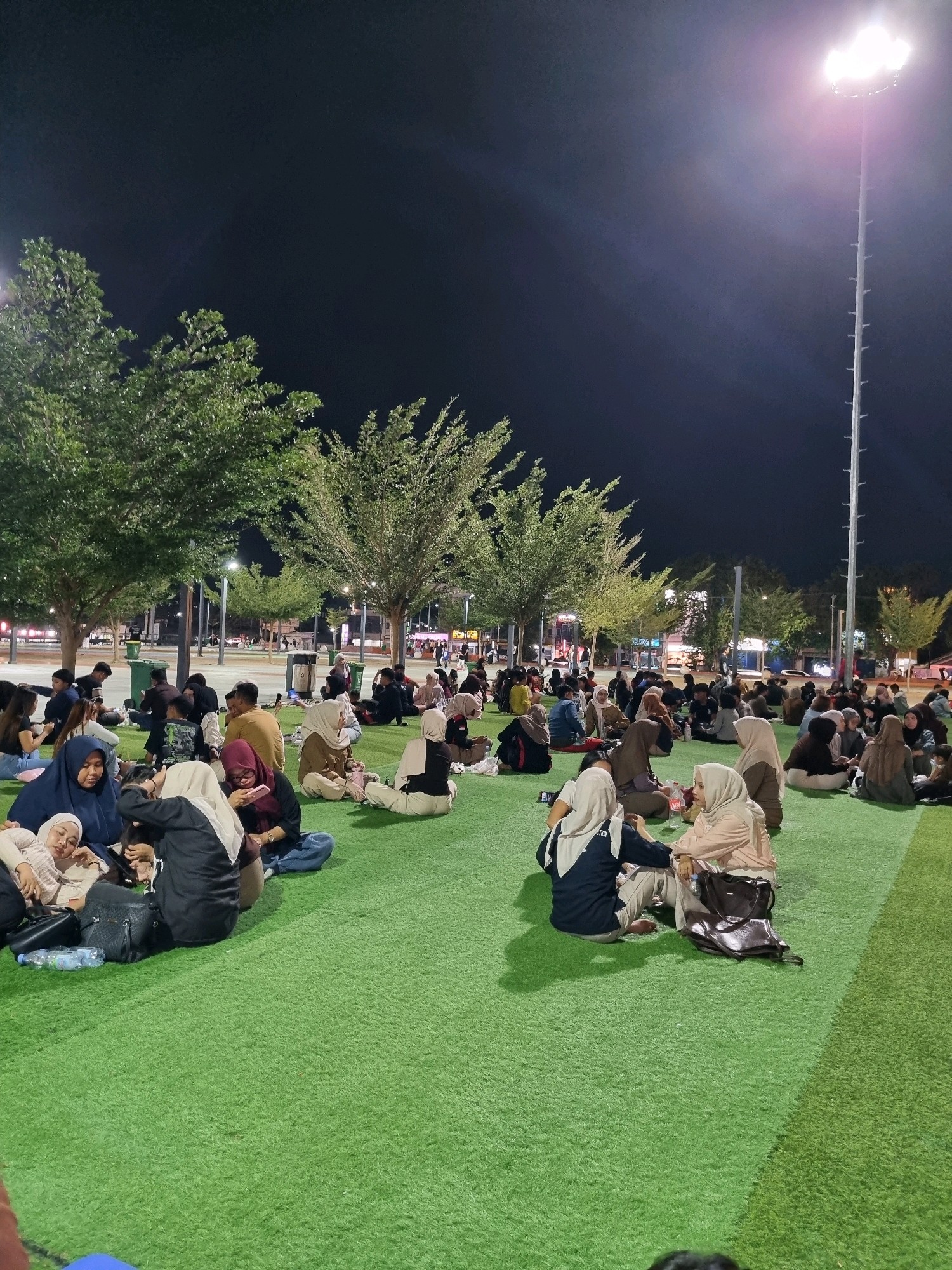 An outdoor scene at night features a large gathering of people sitting on a bright green, artificial turf area. The area is illuminated by tall, bright stadium-style lights positioned on poles to the right of the frame. Several mature trees with green foliage are scattered throughout the scene, some with artificial lighting beneath them. In the background, there are buildings with illuminated windows, suggesting an urban setting.
The people, predominantly dressed in casual attire and many wearing headscarves, are seated in groups on the grass, facing each other or looking towards a common point. Some are engaged in conversation, while others appear to be resting or observing. Various objects are visible on the ground, including bags, water bottles, and personal belongings. The overall atmosphere appears relaxed and communal. The specific location is identified as Tanamodindi, Indonesia.