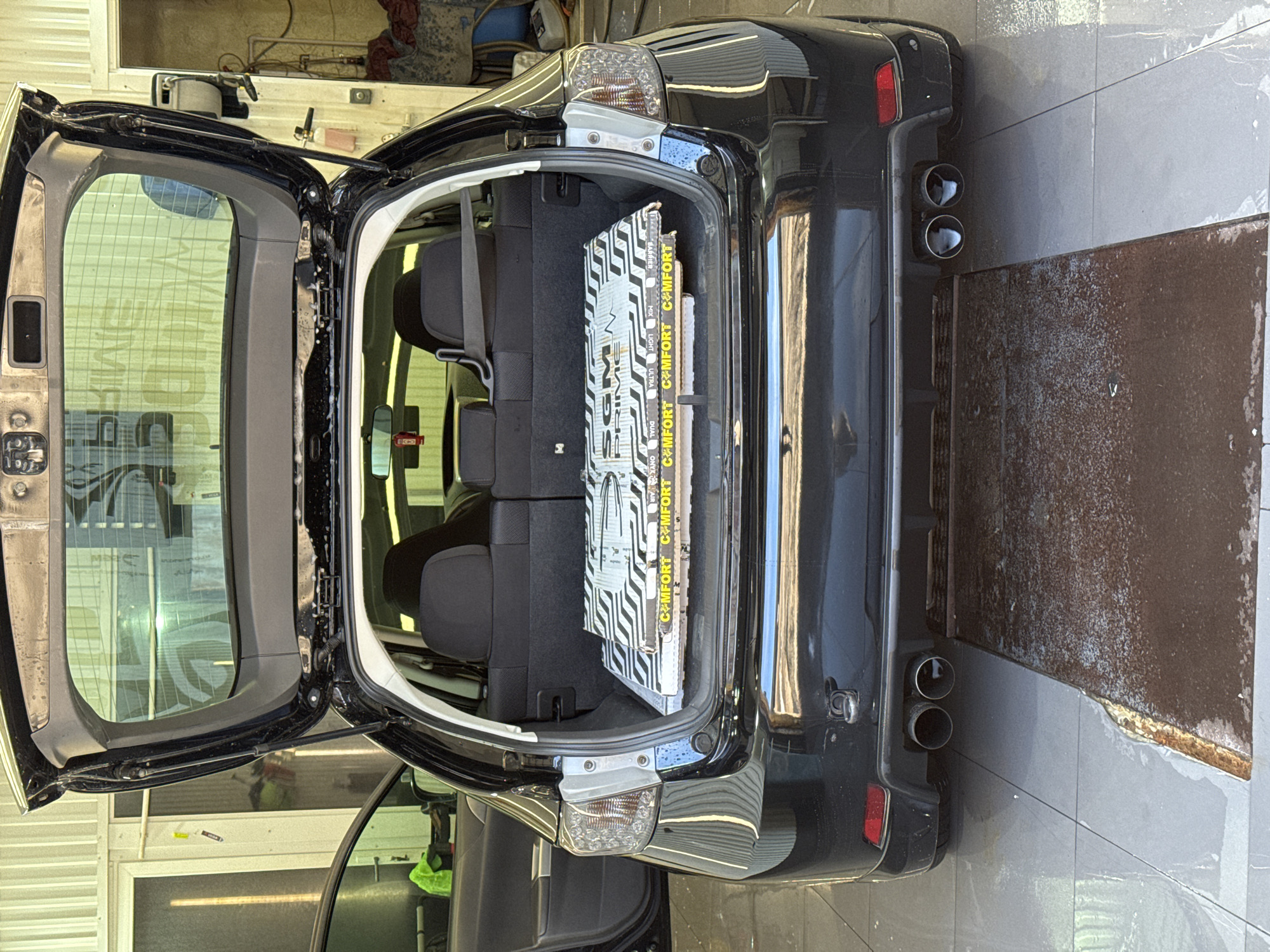 The image shows the open trunk of a black hatchback car, likely a Subaru Impreza, filled with soundproofing material. The trunk contains two boxes of soundproofing; one is branded "SGM Prime", the other "Comfort". The vehicle is in what appears to be a car wash or detailing garage. The location is likely Chaikovsky, Russia. The scene is indoors, illuminated by artificial lighting. No people are directly visible. The floor is tiled and wet. The car's exhaust pipes are visible on the lower right of the image, adjacent to a metallic ramp.