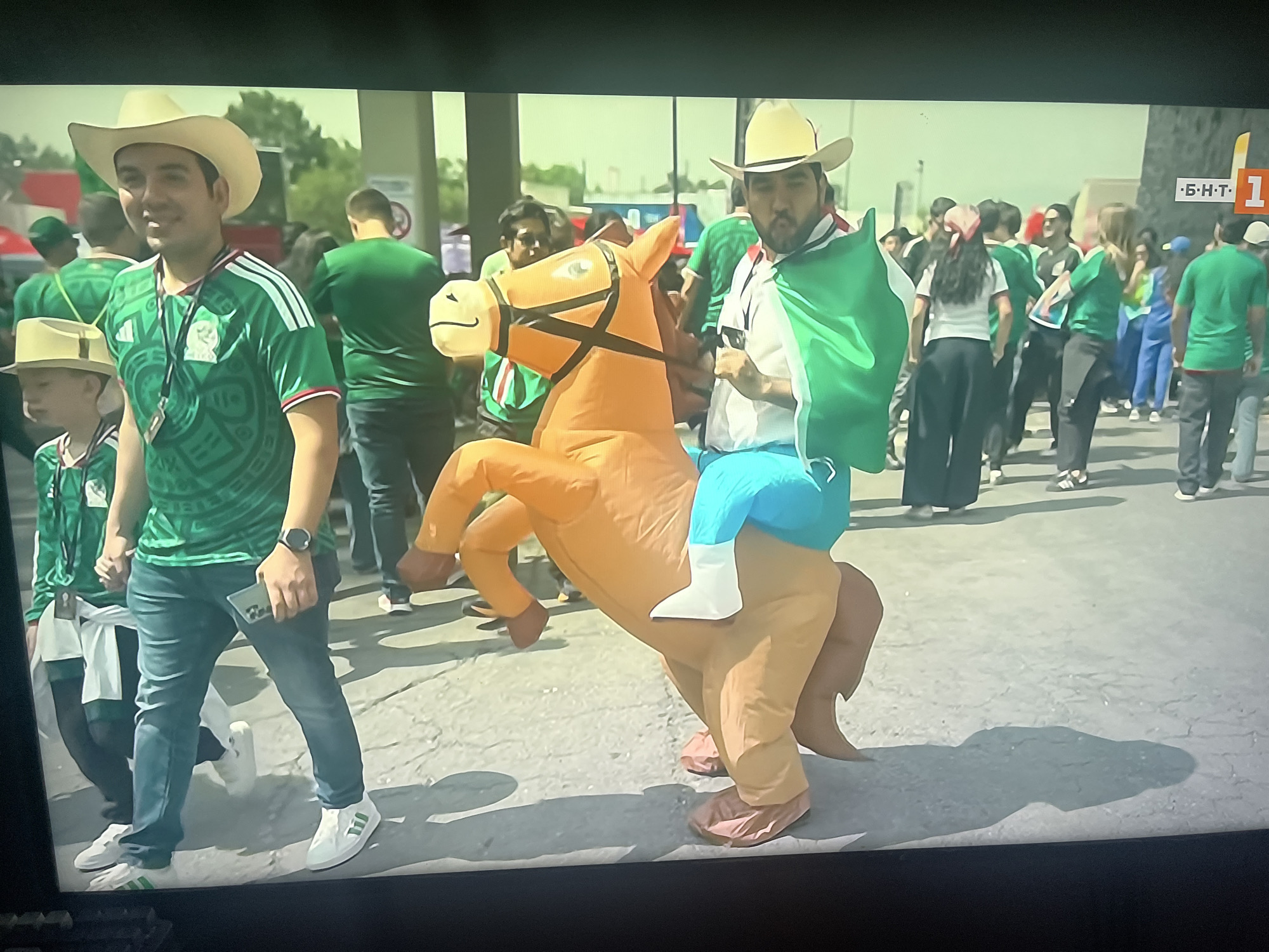 In the city of Ruse, Bulgaria, an outdoor scene unfolds with a group of people, many wearing green Mexican national team jerseys and cowboy hats, indicating a possible sporting event or celebration. A man in the foreground, sporting a white cowboy hat and a green jersey with an Aztec pattern, is holding a mobile phone. To his right, another man is wearing an inflatable horse costume in a bright orange color, with blue pants and white boots. He is draped with a green and white flag, likely representing Mexico. The man in the costume is holding the flag with his right hand and the horse's reins with his left. Behind them, a crowd of individuals, predominantly in green attire, are visible, suggesting a festive atmosphere. The ground is paved and shows cracks, with shadows cast by the people and the inflatable horse, indicating a sunny day. The background features a building with a white sign bearing Cyrillic letters and the number "1", suggesting a commercial or administrative area in Ruse.
