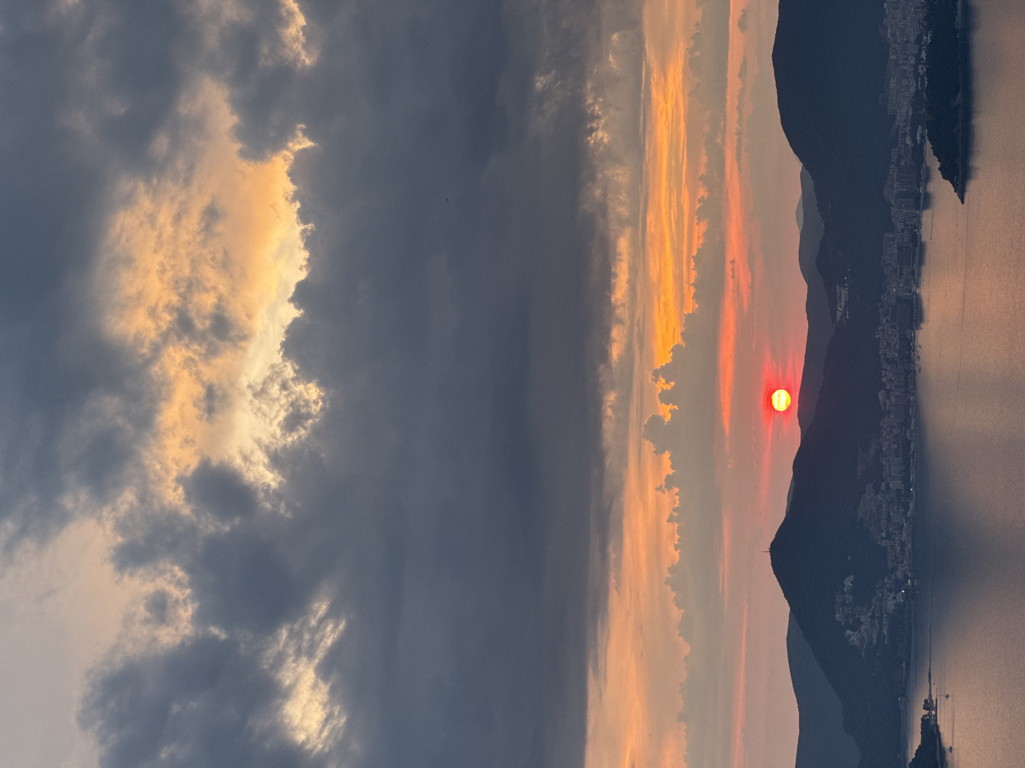 The image captures a dramatic sunset over a coastal city, likely Sveti Stefan, Montenegro. A vivid red sun is partially obscured by the silhouette of a large, dark mountain peak in the mid-ground. The mountain is dotted with buildings, suggesting a town or city nestled on its slopes. The water in the foreground is calm and reflects the warm colors of the sky. The sky itself is a spectacle of color, with layers of dark, heavy clouds in the upper portion contrasting with vibrant oranges and yellows closer to the horizon. Smaller, lighter clouds are scattered throughout, catching the last rays of the setting sun. The overall impression is one of natural beauty and a serene end to the day, with a touch of dramatic intensity from the darkening clouds and the brightly colored sun. There are no visible people in the image.