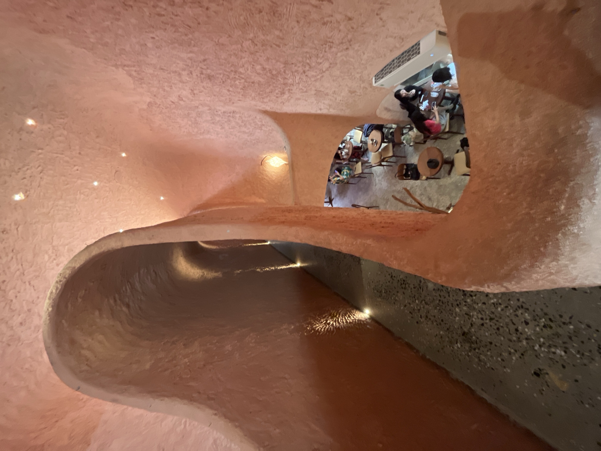 This image captures the interior of the Aurora restaurant in Nha Trang, Vietnam, showcasing its distinctive organic, cave-like architecture with textured pink walls. The perspective is from a higher vantage point, looking down into the dining area and along a curving walkway.
In the foreground and midground, the smooth, undulating pink walls dominate, creating a sense of enclosed, earthy spaces. The walkway, visible on the left, is paved with speckled grey concrete and illuminated by small, recessed lights along its base, creating a gentle glow.
The dining area below features several round tables with wooden chairs, occupied by patrons. On the right side of the image, a group of young women are seated at a table, engaged in conversation and looking at their phones. Another table with two individuals is partially visible further back. The overall ambiance is warm and intimate, enhanced by soft, ambient lighting, including a diamond-shaped lamp hanging from the ceiling. An air conditioning unit is visible mounted on the wall near the seated patrons. The rough texture of the walls suggests a deliberate artistic choice, contributing to the restaurant's unique aesthetic.