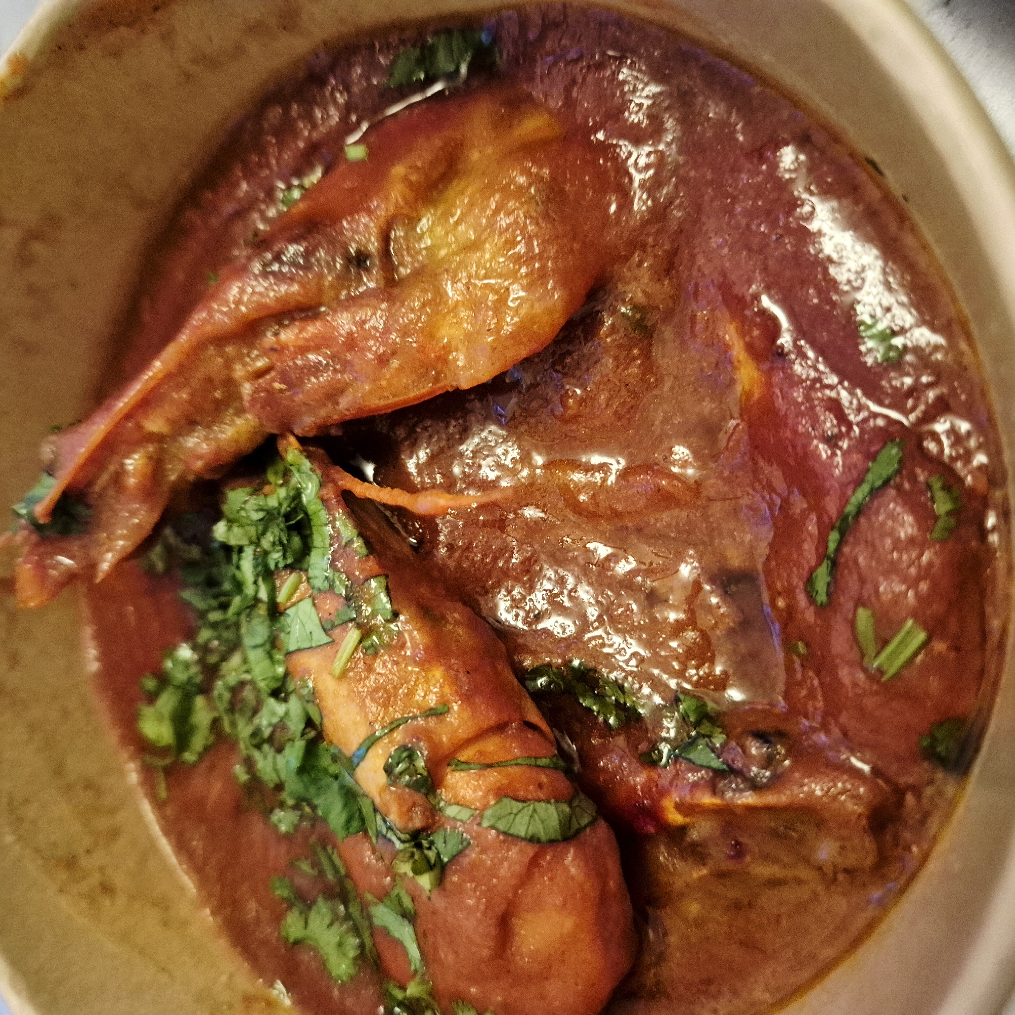 The image shows a bowl of what appears to be shrimp curry, garnished with cilantro, in Strasbourg, France. The dish is contained within a round bowl with a tan or beige exterior. The curry is a rich, reddish-brown color and has a glossy sheen, suggesting it is freshly prepared. At least three large shrimp are visible in the curry, with the tails protruding towards the center. The cilantro is scattered across the shrimp and the surface of the curry. The setting implies an indoor environment, with the focus being exclusively on the presentation of the food.