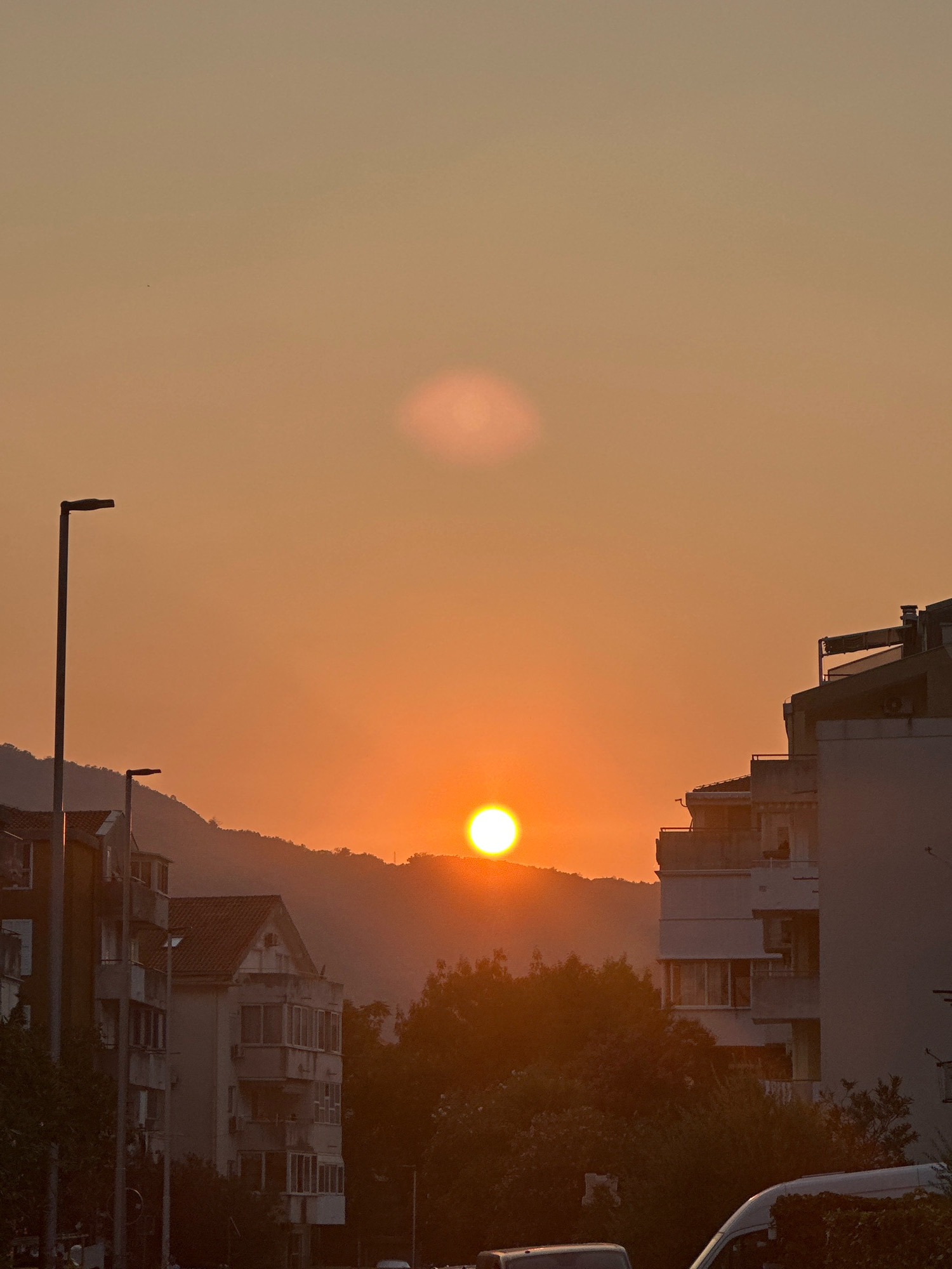 The image captures a sunset in Budva, Montenegro. A bright orange sun is positioned low in the sky, partially obscured by the silhouette of a mountain range in the background. The sky is a gradient of warm oranges and yellows, with a faint, lighter cloud formation visible above the sun. In the foreground, modern apartment buildings with balconies line the street. To the left, a tall street lamp stands sentinel. Trees with dark green foliage create a dense layer between the buildings and the mountain. The presence of vehicles, including a white van, suggests an urban setting. The overall impression is one of a peaceful evening scene in a coastal city.