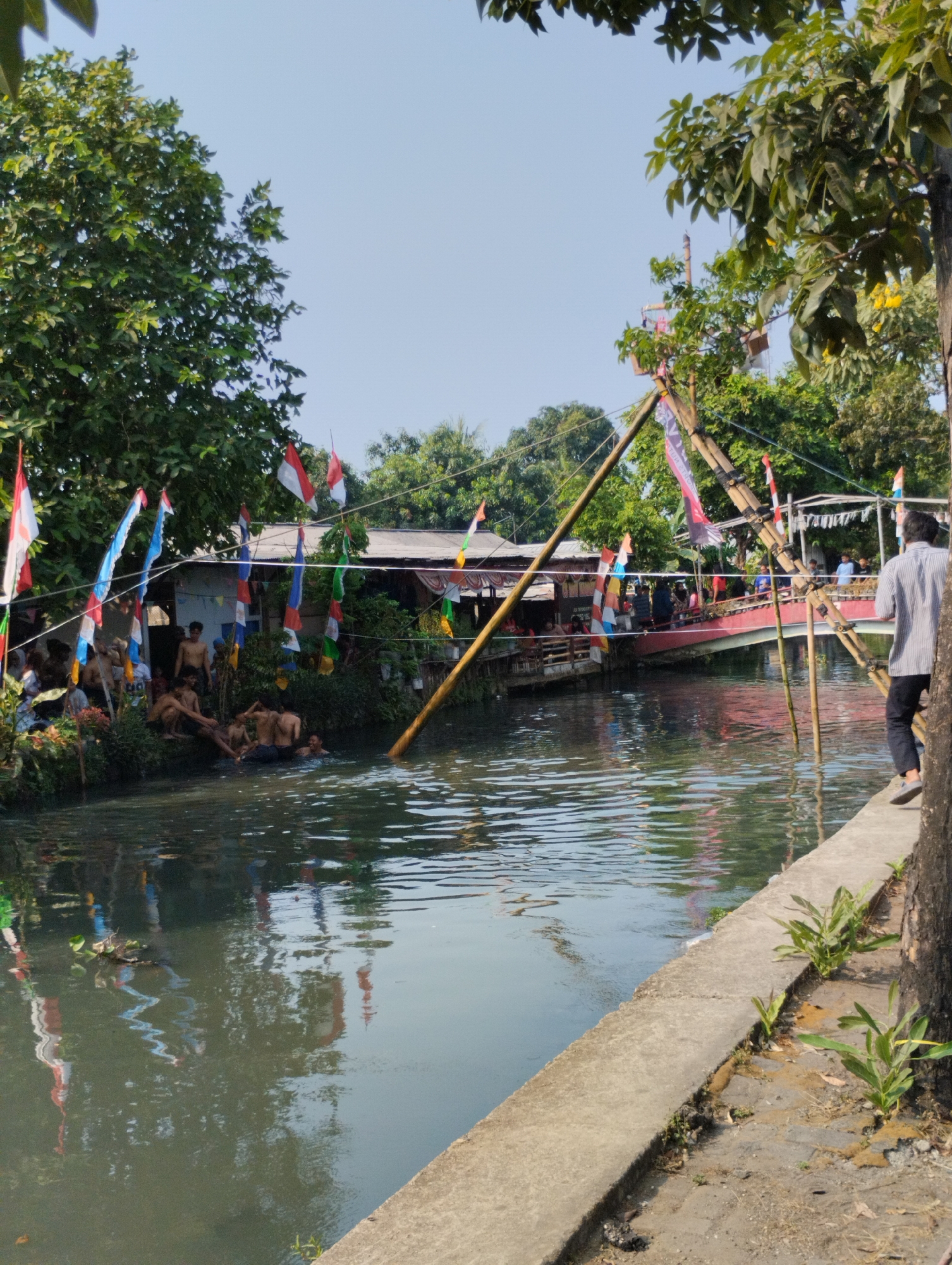 In Bekasi, Indonesia, a group of young men are gathered by the edge of a murky canal, some wading in the water and others sitting on the bank. Indonesian flags are strung across the canal, fluttering in the breeze, hinting at a festive occasion or a commemoration. A red bridge arches over the canal in the background, with more buildings and greenery visible beyond. The scene is bathed in sunlight, casting reflections on the water's surface and highlighting the lush trees that line the canal. The overall atmosphere appears relaxed and communal, with the canal serving as a central gathering point for the community.