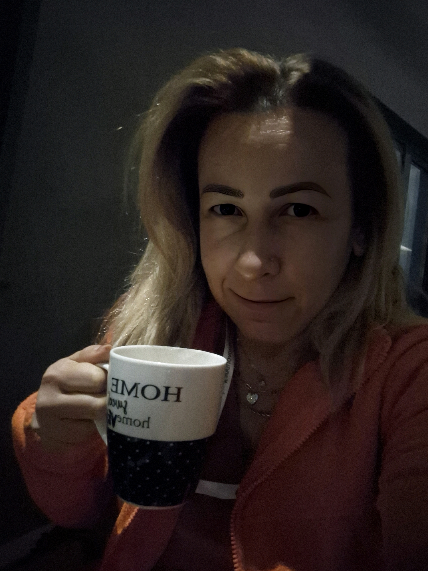 A woman is shown from the chest up, holding a white mug with black polka dots and text that reads "HOME" and "POWER". She has blonde hair and is wearing an orange zip-up hoodie. The background is dark and out of focus, suggesting an indoor setting with low lighting.