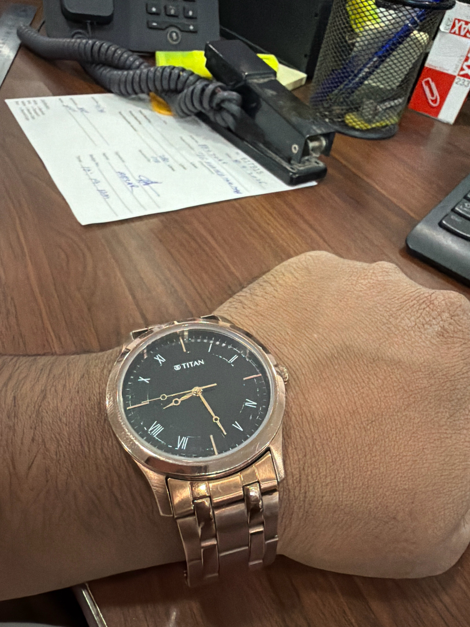 A close-up, eye-level shot shows a rose gold Titan watch on a person's wrist, resting on a wooden desk. The watch has a black dial with Roman numerals and gold hands, indicating the time is approximately 10:10. In the background, a portion of a black office phone with a coiled cord, a stapler, a metal mesh pen holder filled with pens and markers, and a white piece of paper with handwritten text are visible. A sliver of a keyboard can be seen on the right side of the frame. The scene appears to be an office environment. No specific location in Riffa, Bahrain, is identifiable from the image.