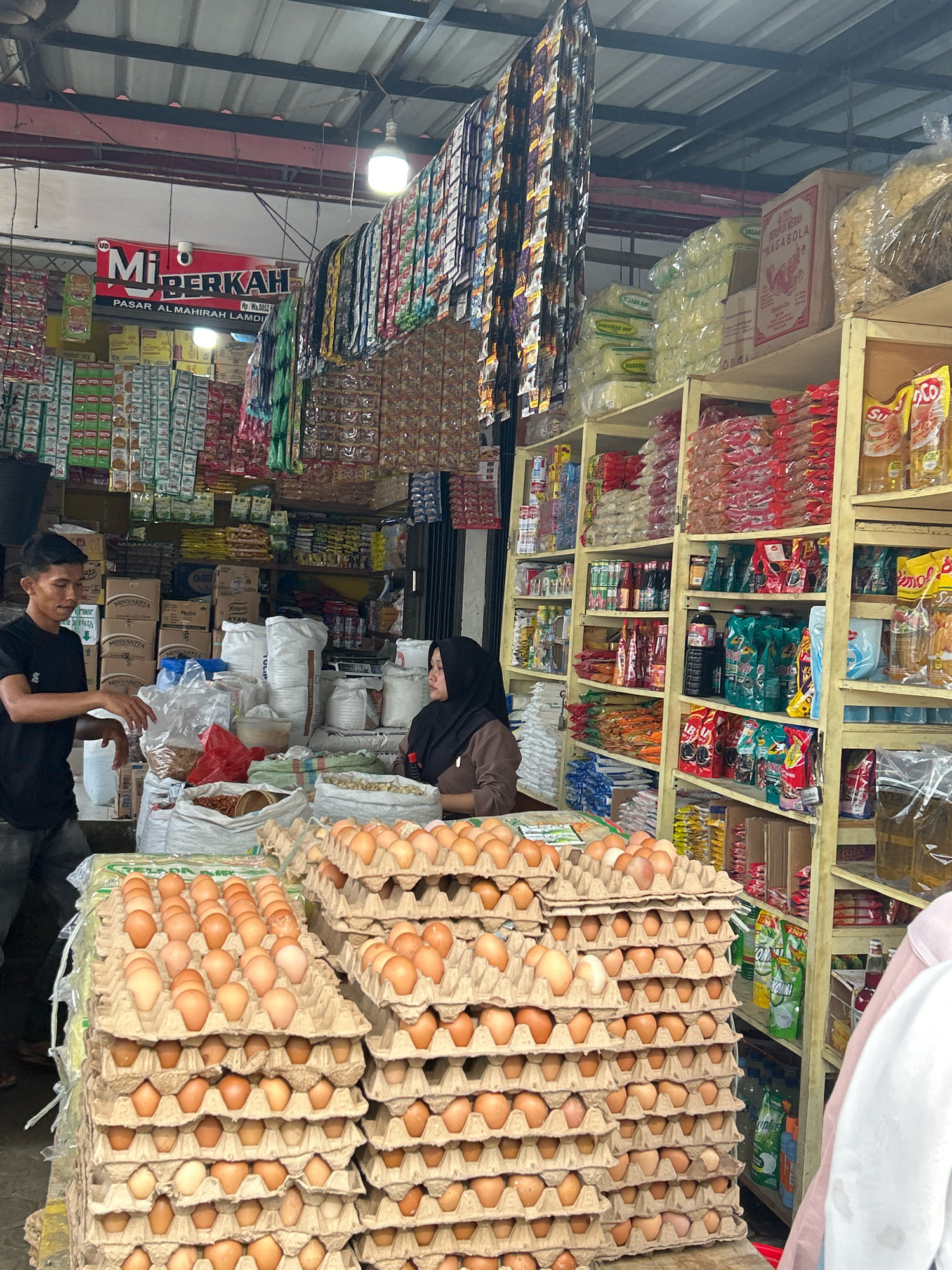 This is an eye-level medium shot of a food stall in Banda Aceh, Indonesia, likely a traditional market. The stall is filled with a variety of goods, including stacked cartons of eggs in the foreground, bags of what appear to be rice or flour, and numerous packaged food items on shelves in the background.
Two individuals are present: a man in a black t-shirt on the left, gesturing with his right hand as if speaking, and a woman wearing a dark hijab on the right, facing the man and appearing to listen. They are positioned behind a counter or table laden with products.
The environment is a covered open-air market, indicated by the corrugated metal roof structure visible at the top of the frame. Various hanging items, likely snacks or spices, are suspended from the ceiling. The lighting appears to be a mix of natural light and overhead artificial lights, suggesting it is daytime. The overall impression is that of a busy commercial setting focused on the sale of staple food items and groceries.