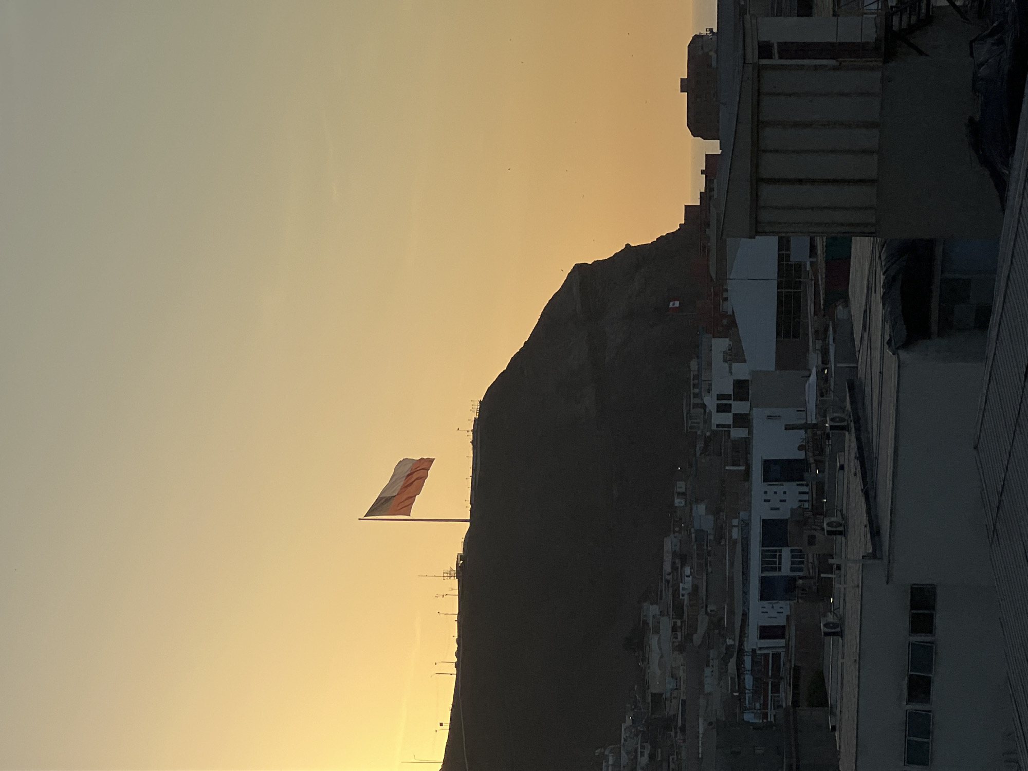 The image shows a landscape at sunset or sunrise. In the foreground, a large flag with horizontal bands of red, white, and possibly another color, is waving atop a tall flagpole. The flagpole is positioned on a ridge or hill, behind which a larger mountain looms. In the middle ground, a dense urban area with numerous buildings appears to be climbing the slope of the mountain. The buildings are predominantly light-colored, with visible windows and air conditioning units. To the right, more modern buildings are visible. The sky is a gradient of orange near the horizon to a light yellow higher up, indicating either the beginning or end of the day. The overall scene conveys a sense of a vibrant urban landscape against the backdrop of a natural mountainous environment. This scene is likely the location in the city of Arica, Chile, where the flag is known as the "Flag of Peace".