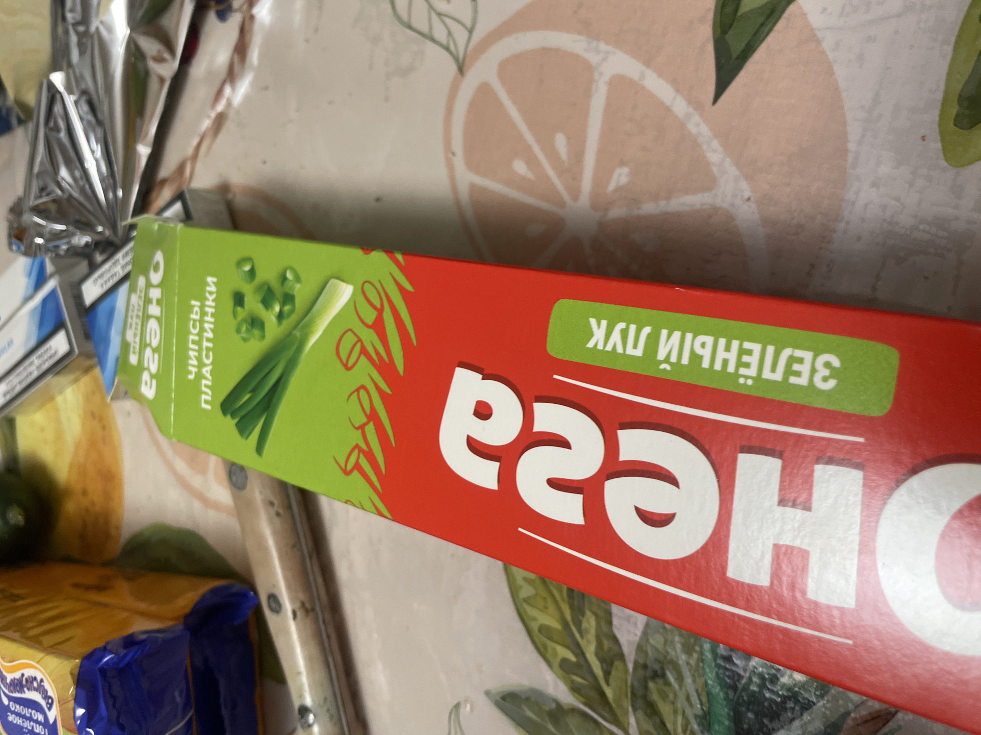 An overhead, close-up shot shows a red and green box of potato chips with green onion flavoring. The box is positioned diagonally across the frame. Text on the box reads "CHEESE" in large white letters and "ЗЕЛЁНЫЙ ЛУК" (green onion) in white letters on a green background. The chips are described as "чипсы пластинки" (chip slices). The background is a table with a pattern of sliced citrus fruit. In the upper left corner, a portion of a yellow package and a blue wrapper are visible. A metal knife with a wooden handle lies horizontally across the lower left portion of the frame. There are no people or animals in the image. The setting is indoors, likely a kitchen or dining area. There is no information to suggest this image is related to the city of Laishevo, Russia.