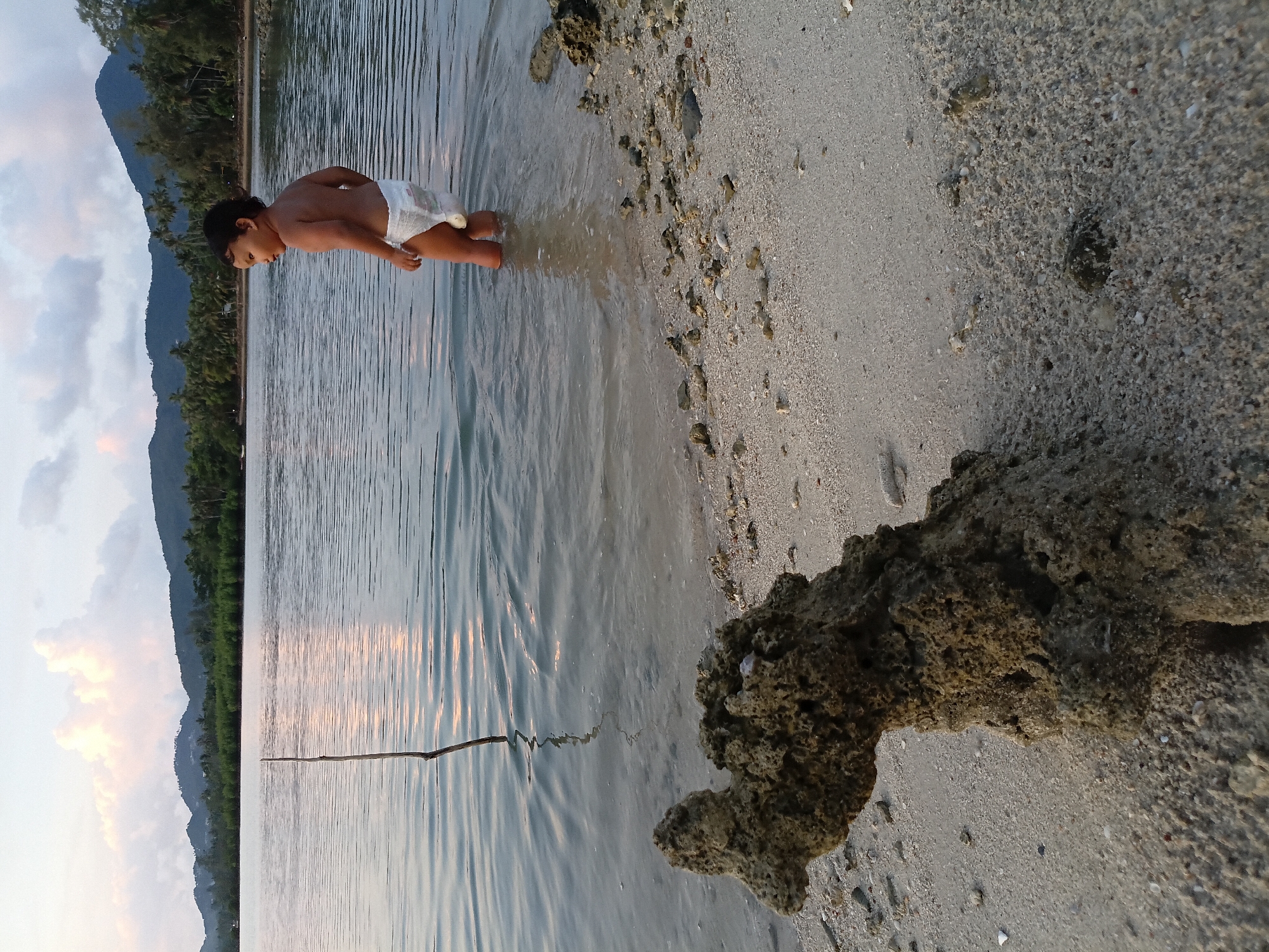 A child is standing in shallow water at a beach in Baan Wok Tum, Thailand. The child, wearing a diaper, is looking down at the water. The water is calm with small ripples. The shoreline is composed of sand and rocks. In the background, there is a mountain covered in vegetation. The sky is cloudy.