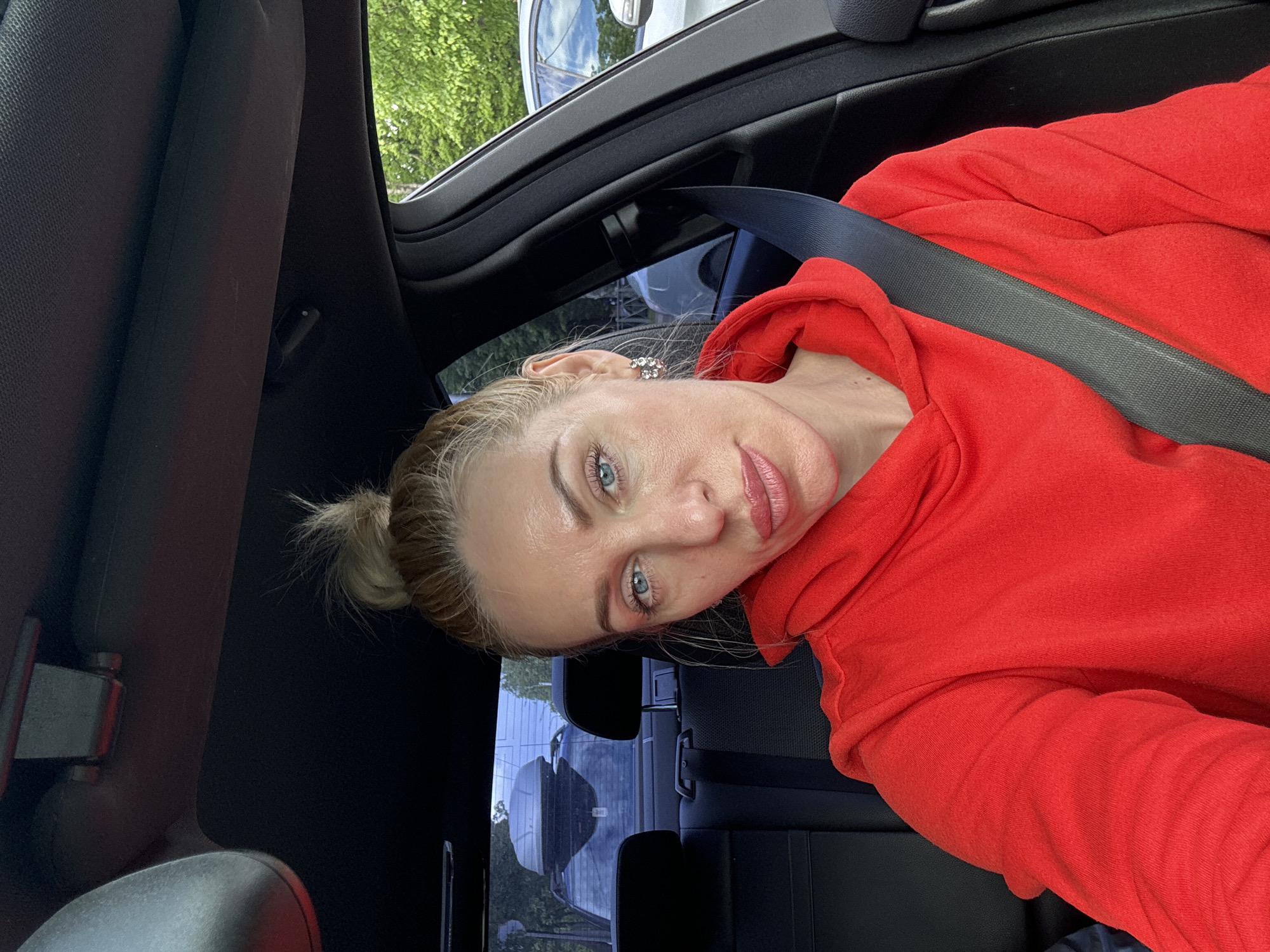 A woman with blonde hair styled in a bun, wearing a bright red hooded sweatshirt, is seen inside a vehicle. She is wearing a seatbelt. The interior of the vehicle is black. Trees and other vehicles are visible through the car windows.