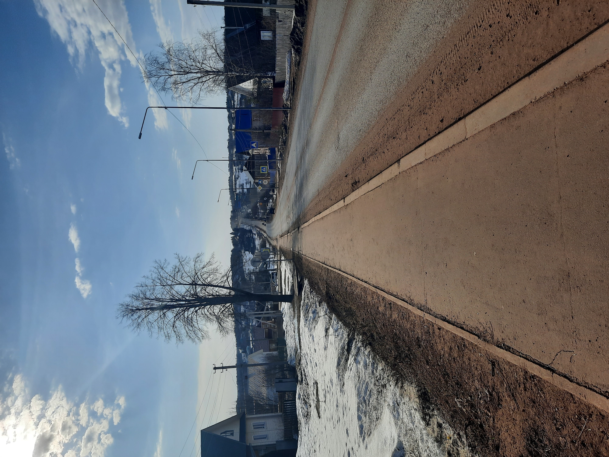 The image depicts a street scene in Krasnogorskoye, Russia. The road is asphalted and slightly wet, stretching into the distance towards a gentle rise in the landscape. A concrete curb separates the road from a strip of dirt and snow-covered ground. Houses are visible on both sides of the road, receding into the distance. Power lines and streetlights line the street. The sky is blue with scattered clouds. A bare tree stands along the roadside. The overall scene suggests a rural or suburban setting on a clear day with remnants of winter.
