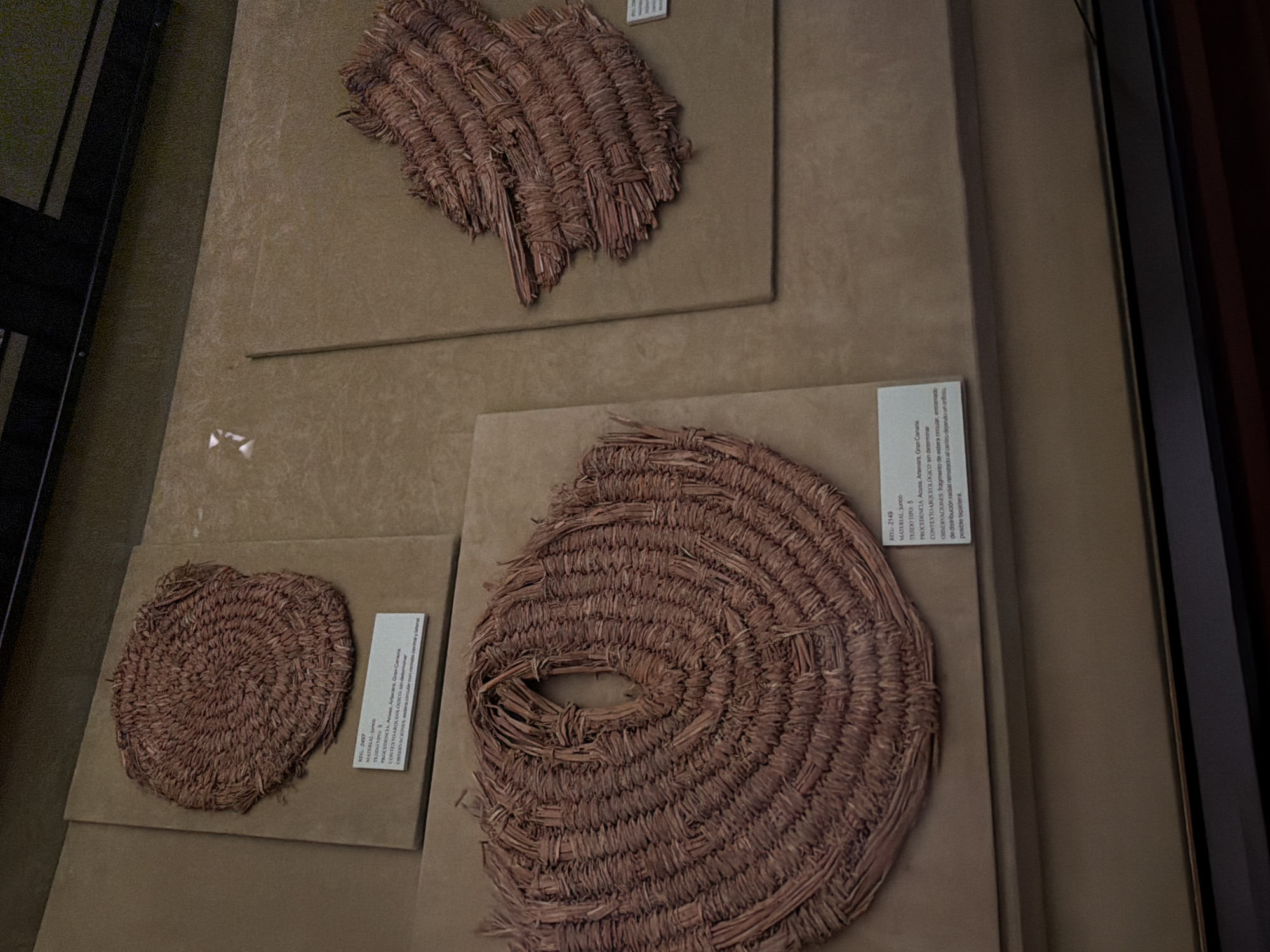 The image shows a museum display case featuring three woven artifacts made of dried plant material, likely reeds or grasses. The display case is divided into sections. On the left, there are two artifacts. The upper artifact is a circular, coiled mat with a textured surface. Below it, a larger, oval-shaped artifact with a central opening is prominently displayed. Its weave is more open and visibly constructed from bundled plant fibers. To the right, a third artifact, a fragmented piece of woven material, is displayed against a textured background. White descriptive labels are visible in front of the two artifacts on the left. The overall environment appears to be a museum with neutral-toned walls and professional lighting. No people are visible in the frame. The artifacts are likely part of an archaeological or ethnographic exhibition in Las Palmas de Gran Canaria, Spain.