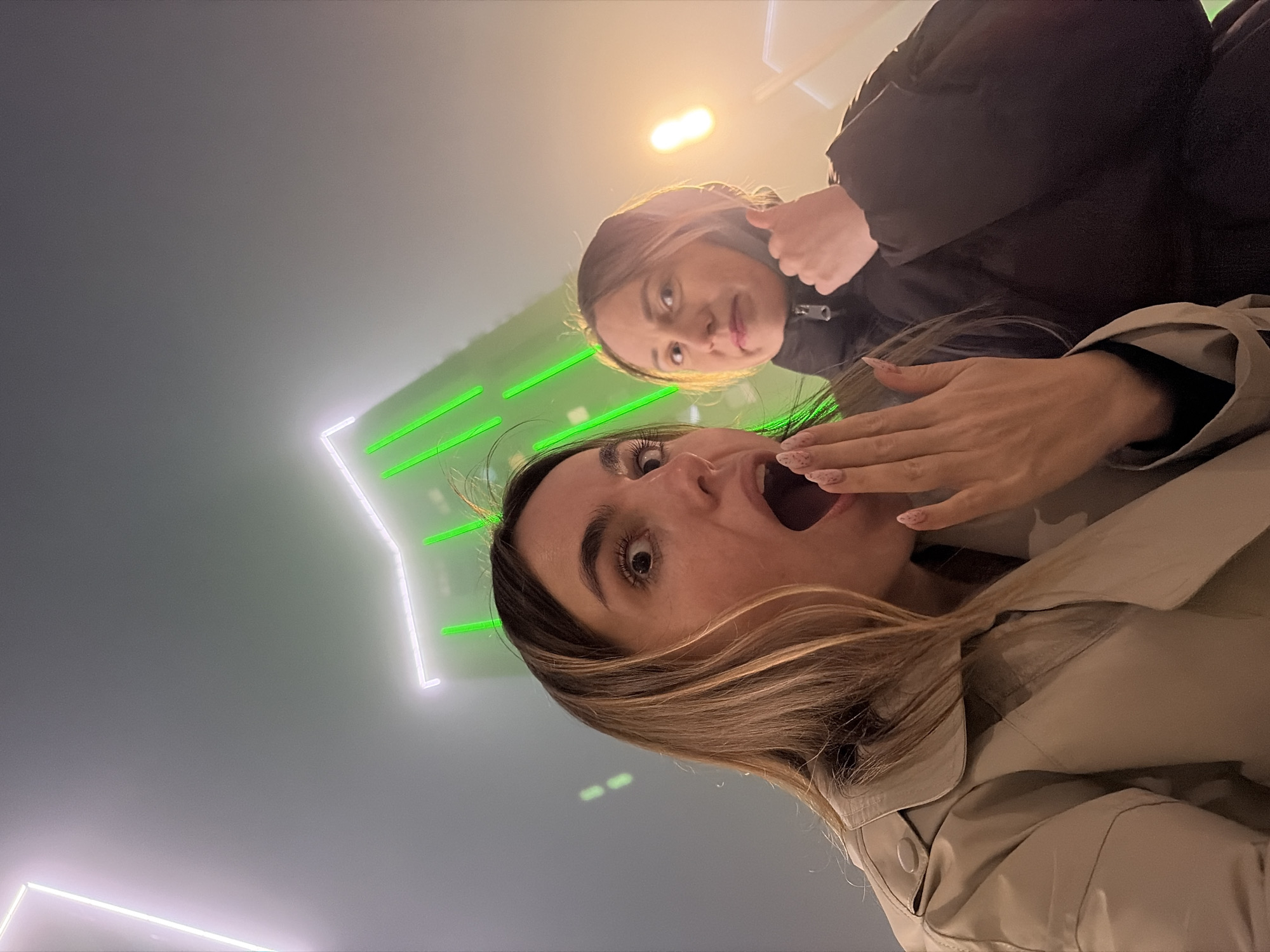 Two young women pose for a selfie in an entertainment venue in Izhevsk, Russia. One woman in a beige trench coat has an open mouth and raises her hand, displaying patterned nails. The other woman in a dark puffer jacket gives a thumbs up. Illuminated neon shapes, including linear polygons and green stripes, create a backdrop. The image has a strong upward angle.