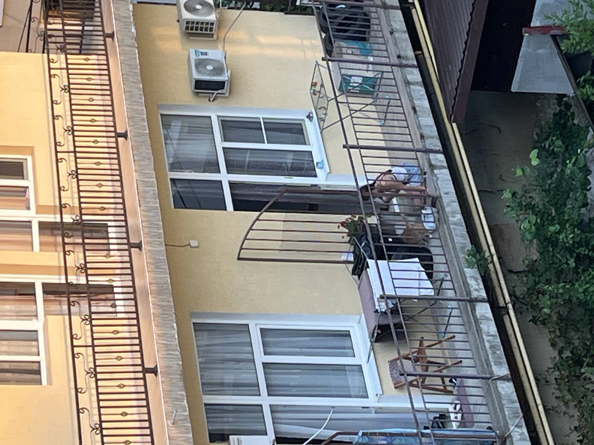 The image depicts a balcony of a building in Sochi, Russia. The balcony is enclosed by a dark metal railing. On the balcony, there are several objects, including a foldable chair, a clothes drying rack with laundry, a table with a game on it, and a large trash bin. Two air conditioning units are mounted on the yellow exterior wall of the building. In the foreground, on the balcony floor, a person is crouching down, and a dog is near them. The person appears to be wearing a tank top and shorts. There are also some plants and greenery visible below the balcony. The overall scene suggests a residential setting with everyday items present on the balcony.