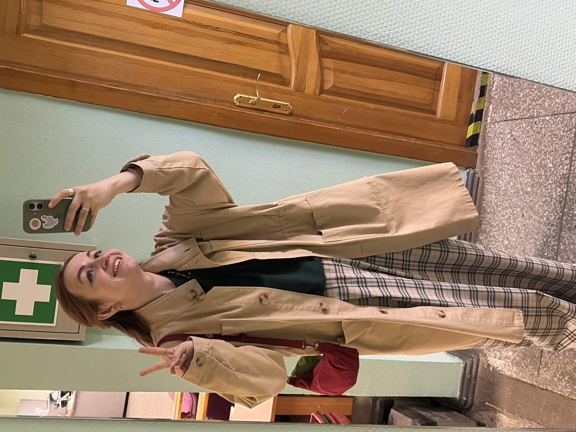 A woman is taking a selfie in a mirror. She is wearing a beige trench coat, a dark green top, and plaid pants. She is holding her phone in her left hand and making a peace sign with her right hand. She is smiling and looking at the phone. The background includes a wooden door with a brass handle, a green wall, a first-aid kit sign, and a tiled floor. The location appears to be in an indoor setting, possibly a hallway. There is no explicit information in the image to definitively confirm the city of Chaikovsky, Russia.