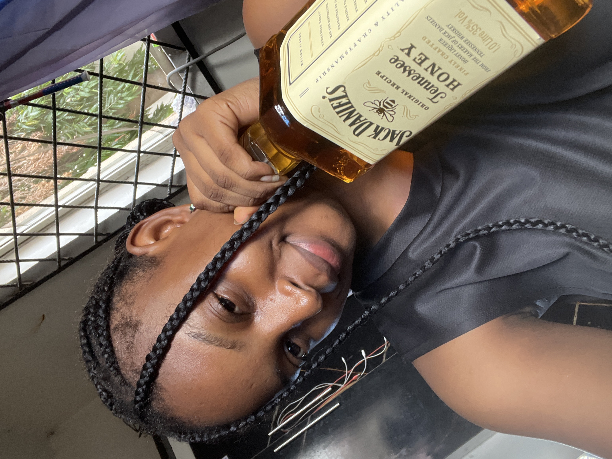 A woman is holding a bottle of Jack Daniel's Tennessee Honey whiskey close to her face. The bottle is partially covering one of her braided hairstyles. The woman is smiling slightly. Behind her is a window with a metal security grill. The setting appears to be indoors. There is no information to place the location in Lokoja, Nigeria.