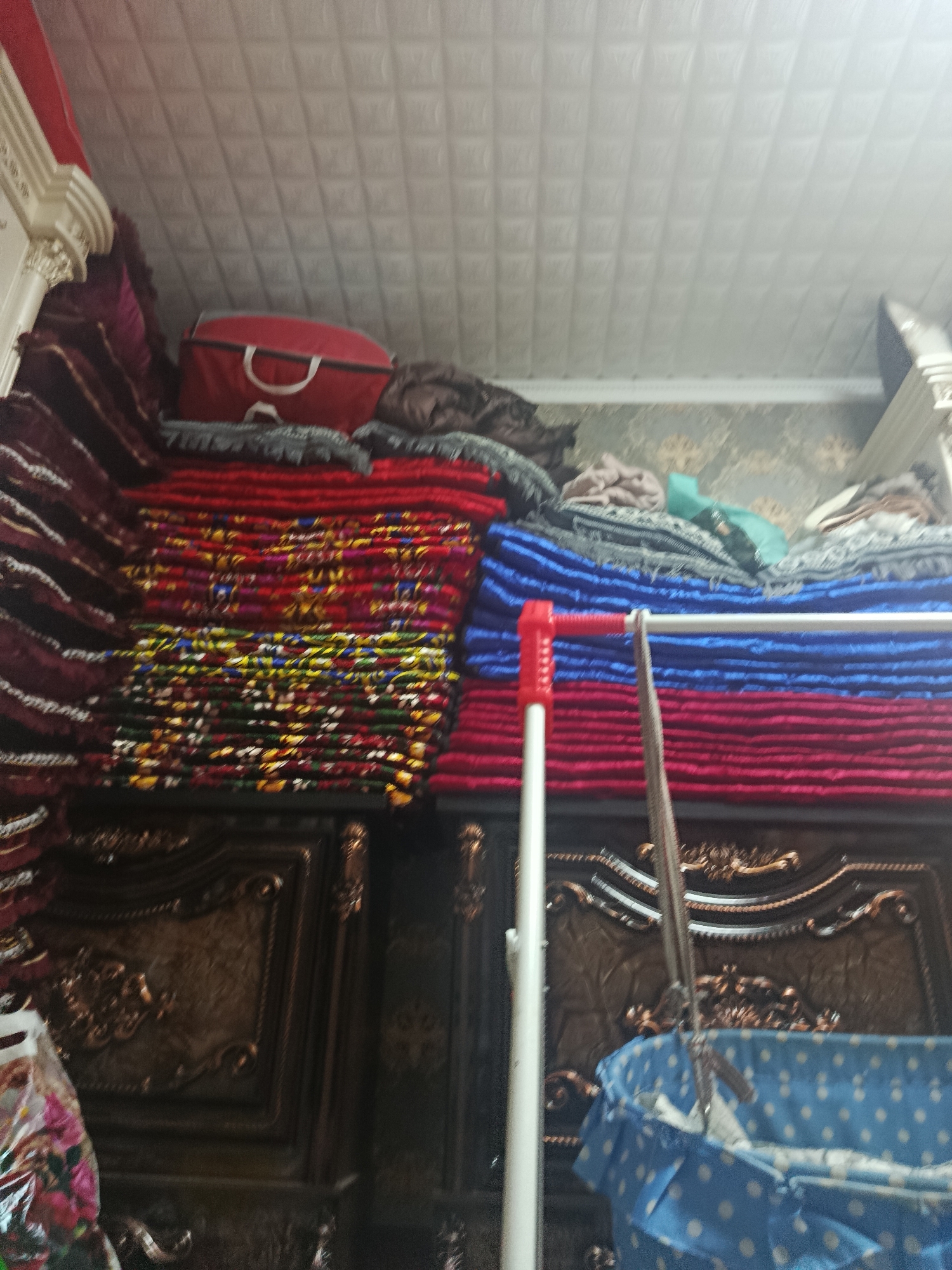 This is a medium shot, eye-level, indoor photograph of a storage area, possibly a closet or a bedroom, with the focus on stacked textiles. In the foreground, a blue and white polka-dotted baby bassinet is partially visible. Behind the bassinet, a white pole with red accents stands vertically. The middle ground is dominated by large stacks of folded fabrics in various colors, including red, blue, and intricate patterns with yellow, green, and pink accents. There are also darker colored textiles, possibly blankets or rugs, stacked on top of some of the colored fabrics. A red duffel bag with white handles rests on the top stack of textiles. The background shows a textured ceiling and a wall with a faint floral pattern. The overall lighting is dim. The scene appears to be in a residential setting in an unknown location.