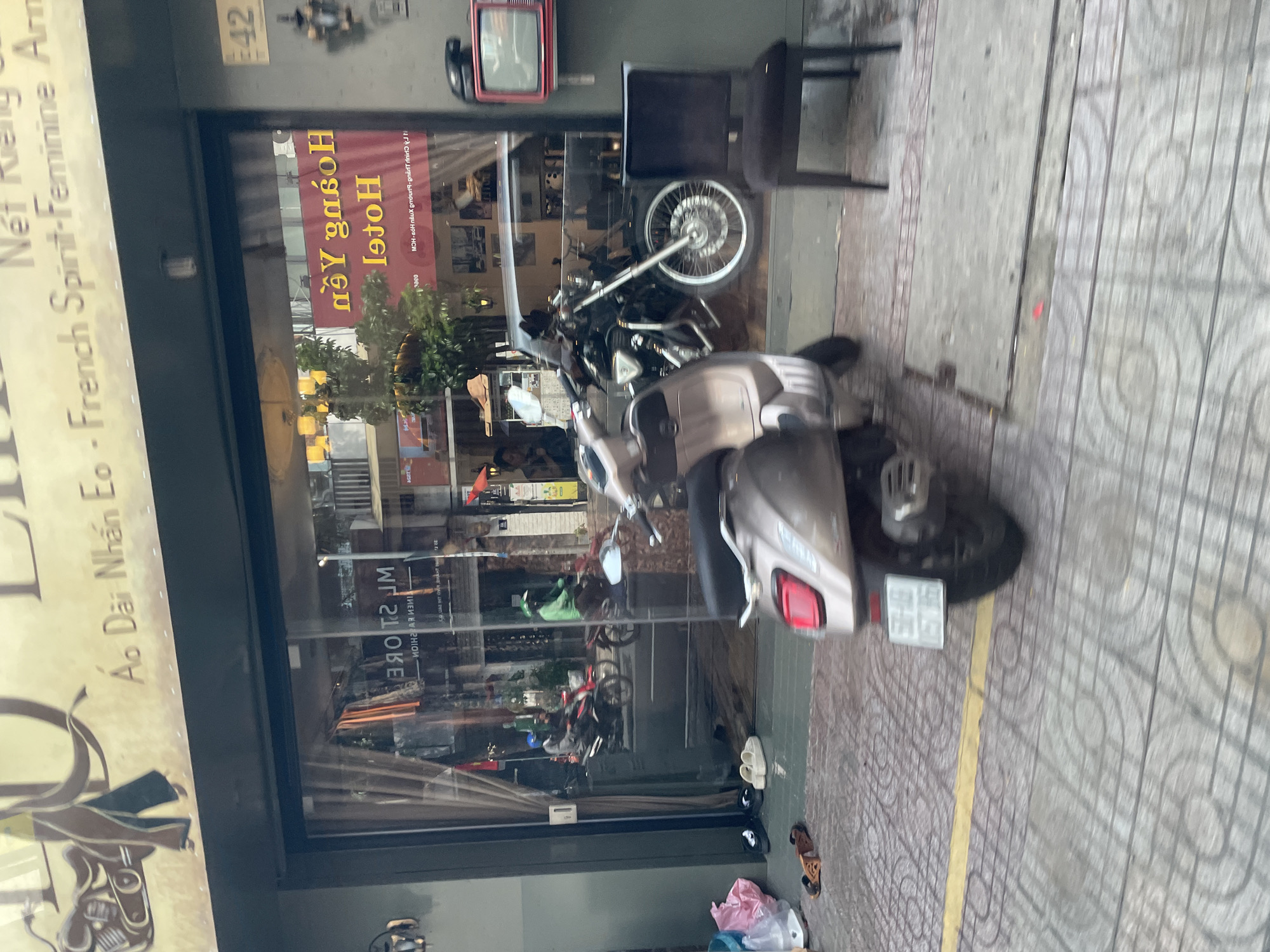 The image shows a storefront in Thủ Đức, Vietnam, with a street view. The storefront has a large glass window displaying motorcycles and interior elements, including a red vintage television mounted on the wall. A beige scooter is parked in front of the storefront, and various footwear is scattered on the tiled sidewalk. The street is paved with interlocking concrete blocks, and a yellow line marks the edge of the sidewalk. Reflections in the window reveal street activity, including people on motorcycles and buildings. The signage above the storefront is in Vietnamese and English, suggesting a commercial establishment, possibly a clothing store or hotel. The overall scene depicts an urban streetscape in Vietnam with elements of daily life and commercial activity.