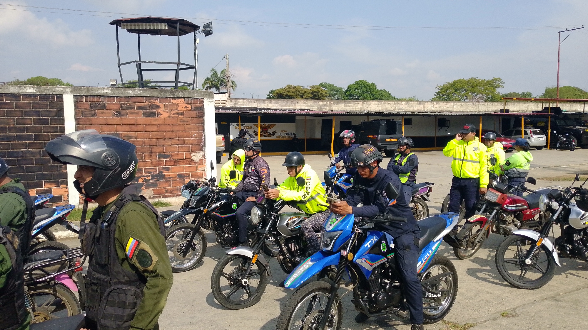 The image captures a gathering of uniformed men, presumably law enforcement or military personnel, with their motorcycles in an outdoor paved area, likely a compound or station, in Tinaquillo, Venezuela. The scene takes place on a bright, sunny day under a clear sky.
The subjects are numerous men, most wearing dark helmets. Their attire varies: some wear olive green/khaki uniforms with black tactical vests, one featuring a Venezuelan flag patch; others are in dark blue uniforms. Many wear bright neon yellow high-visibility jackets or hoodies, some with text like "CONAS" or "C.P.B." printed on them. They are positioned around and on a variety of motorcycles, which include both dirt bike-style and standard street models. Some individuals are actively using their mobile phones, while others are conversing.
In the background, a substantial brick wall separates the foreground from what appears to be a secure facility. A metal watchtower or guard post stands prominently above this wall. Beyond, low-rise buildings with carports house additional vehicles, including cars and a dark tactical-looking truck. A sign on one of the background buildings reads "EL HAMPA NO TIENE" and "PEDRO EL PUEBLO Y SUS GUARDIANES" alongside an emblem. Other visible text includes "EDGE" on a helmet, "C.P.M.T." on another, and "POLICIA" on a motorcycle fender. The overall impression is one of a security force preparing for, or concluding, an operation.