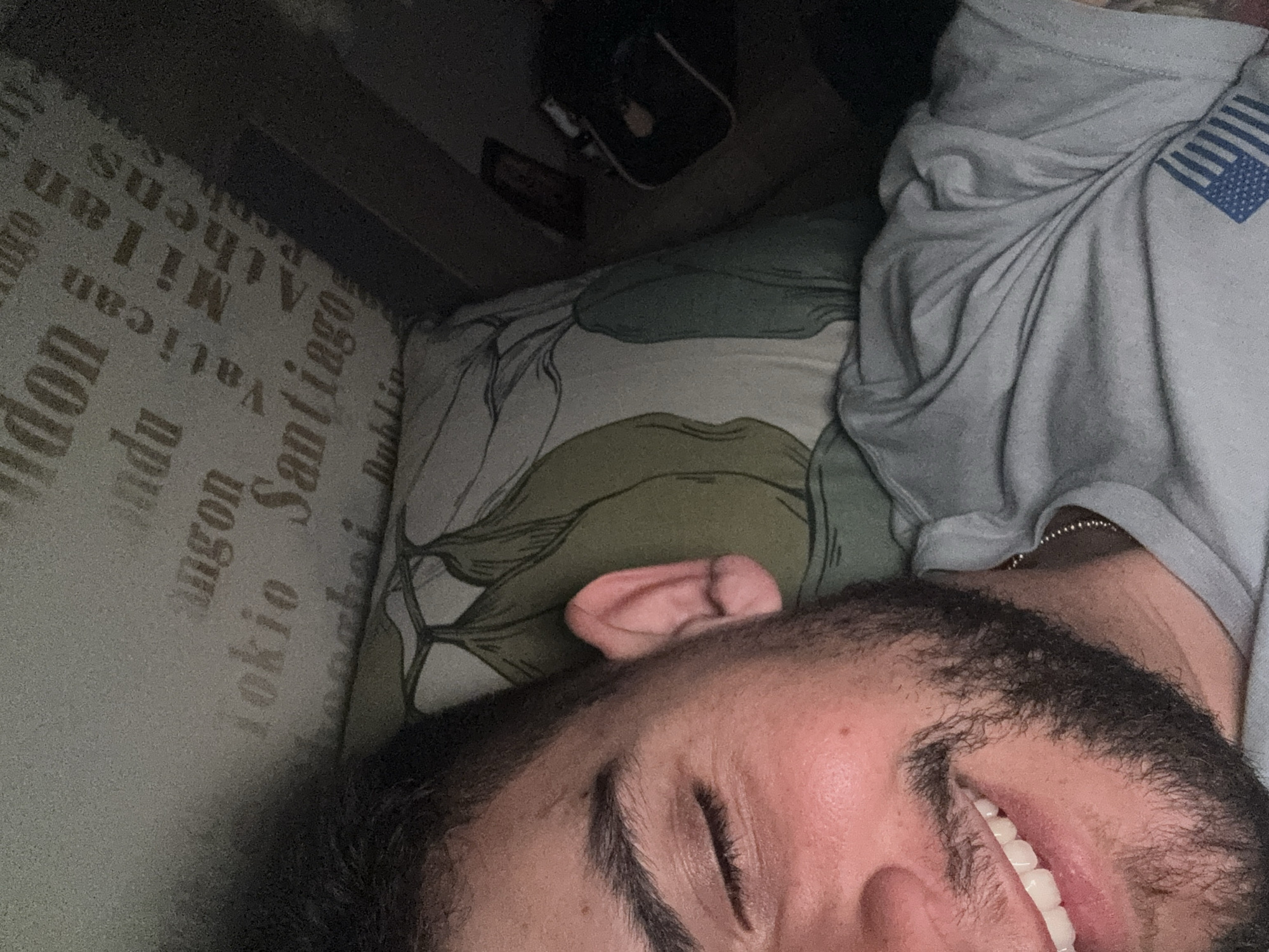 A close-up, rotated image shows a man lying down. He is facing away from the camera and has his eyes closed. He is smiling, and his teeth are visible. He has dark hair and a beard. He is wearing a grey T-shirt with what appears to be a small American flag design on the shoulder. He is lying on a pillow with a floral design on top of a patterned surface featuring city names like "London", "Milan," "Vatican," "Tokyo," "Santiago," and "Dublin." The background is dark, and in the upper portion of the image, there is a dark object that appears to be a small bag or case. The image does not provide information to definitively place the subject in Sofia, Bulgaria.
