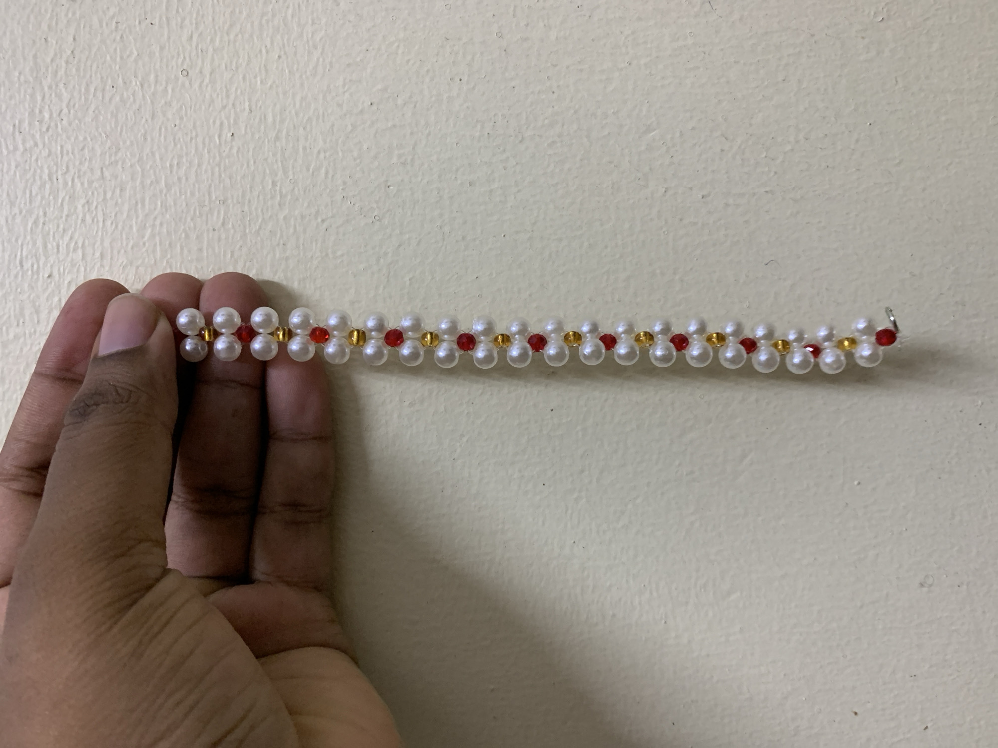 A hand holds a string of pearls with gold and red beads, against a cream-colored wall.