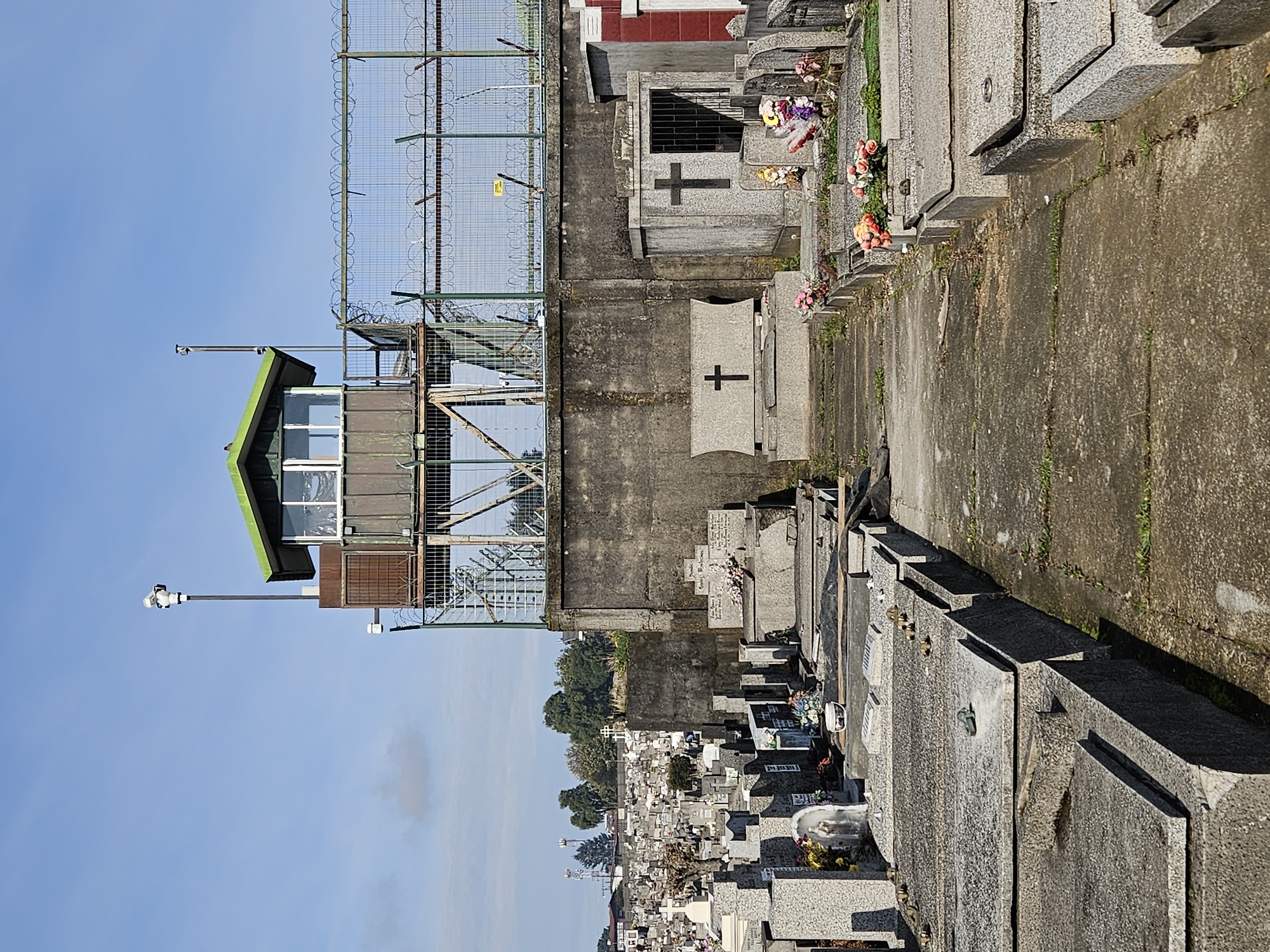 The image shows a cemetery in Osorno, Chile, viewed from an elevated position near a prison watchtower. Rows of gravestones and mausoleums stretch into the distance. Some graves are adorned with flowers. Adjacent to the cemetery is a tall concrete wall topped with a fence and barbed wire, likely belonging to the prison. A watchtower stands next to the wall, equipped with windows, a green roof, and a security camera. The sky is blue and clear.