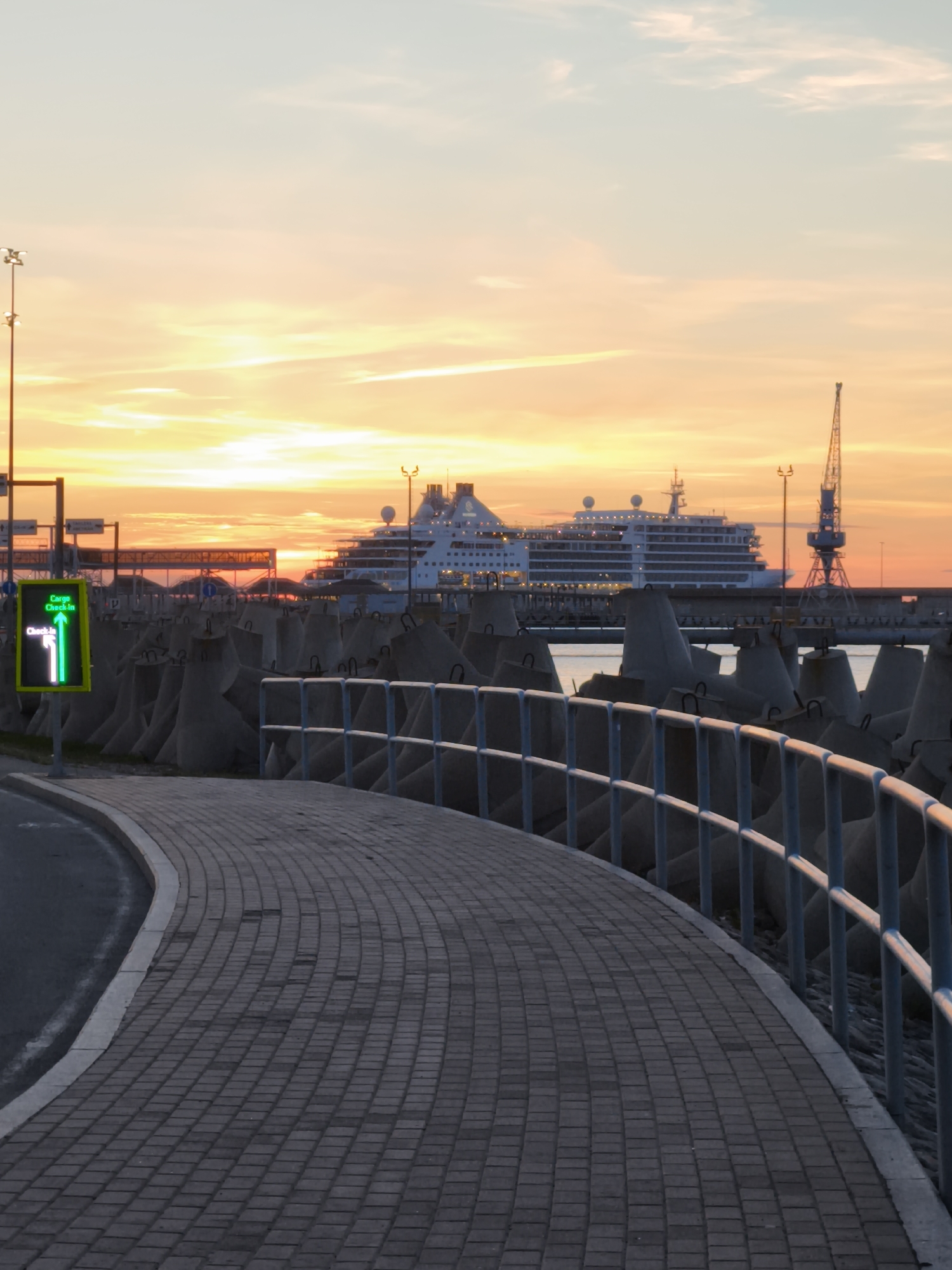 A digital sign displays a green arrow pointing left and the text "Cargo Check-in" in English. The sign is positioned on a paved walkway that curves to the left, with a metal railing to the right. In the background, a large cruise ship is docked at a port, with a crane visible to its right. The sky is filled with the warm hues of a sunset, casting an orange glow over the scene. The location is the Tallinn Port in Tallinn, Estonia.