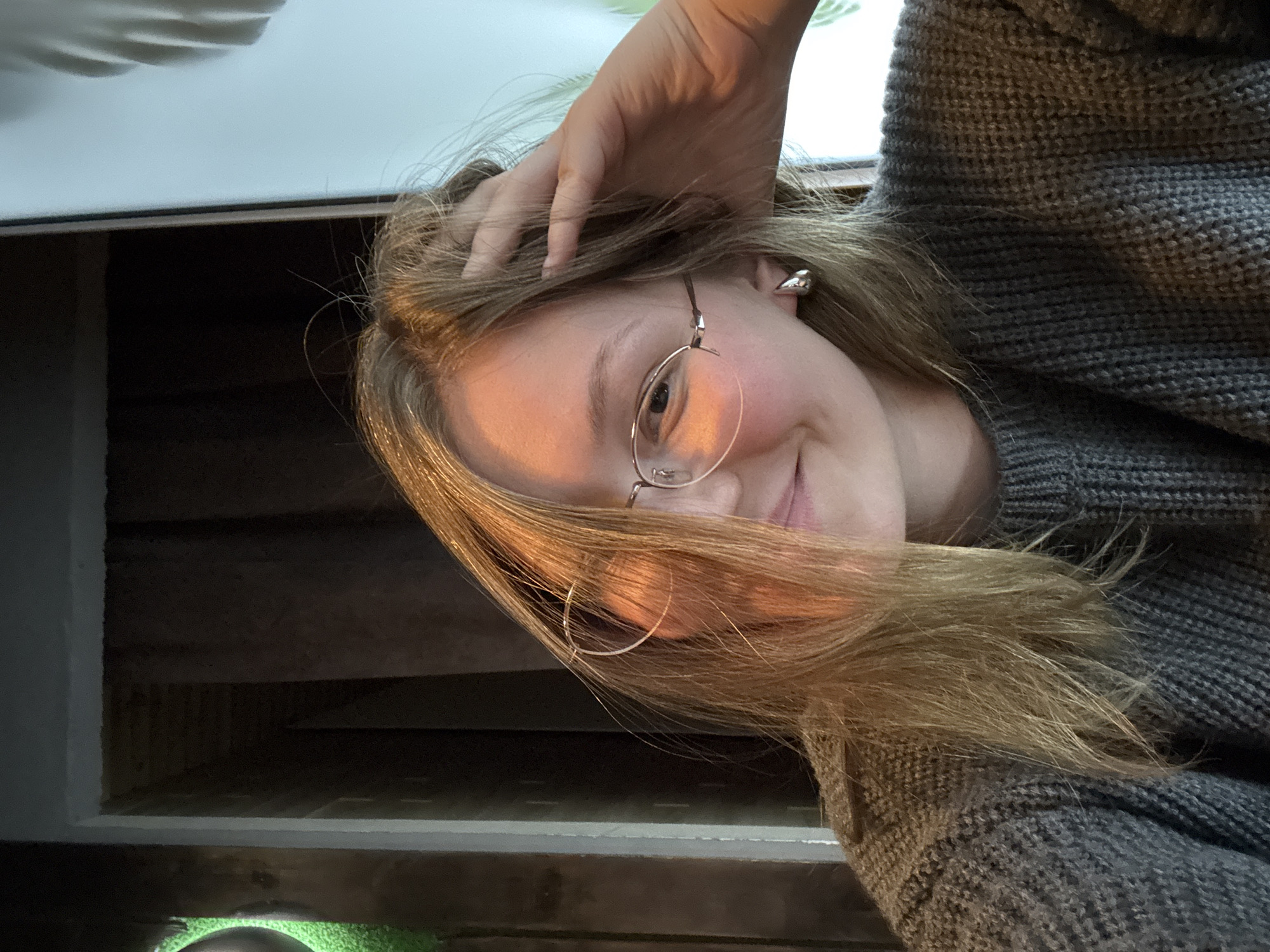 The media file presents a close-up, rotated portrait of a young person, likely female, with long, light brown hair. She is wearing round-rimmed glasses and a grey knitted sweater. Her left hand is casually placed on top of her head, with fingers lightly running through her hair. The subject looks directly at the camera with a soft, relaxed smile.
Notable details include two different earrings: a small, silver, abstractly shaped stud on her left ear, and a thin silver hoop on her right ear. Her fair skin and hair are illuminated by a warm light source, creating gentle highlights and shadows.
The background suggests an indoor setting. Above the subject's head, a light-colored ceiling or wall is visible, with subtle reflected shadows. Below her, there are dimly lit dark wooden structures, possibly shelving units or architectural beams. A vibrant green object or surface, potentially artificial turf or a decorative item, is visible at the very bottom edge of the frame. The image appears to be a personal selfie, conveying a calm and pleasant mood. No text is visible in the image. The photo was taken in Chaikovsky, Russia.