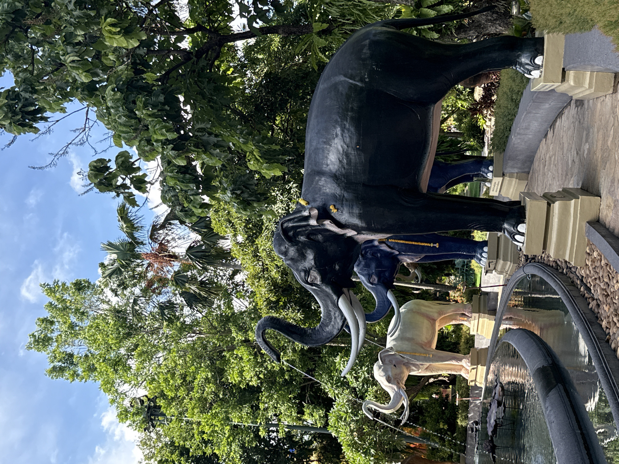 The image depicts a collection of elephant statues in Samrong Nuea Subdistrict Municipality, Thailand. The statues are arranged in a line, with each subsequent elephant smaller than the previous one. The largest elephant is black, followed by a dark blue one, and then two lighter-colored elephants. The statues are positioned on pedestals beside a water feature with a dark, round edge. Lush greenery, including trees with dense foliage, surrounds the statues, with a glimpse of a blue sky and palm trees visible above.