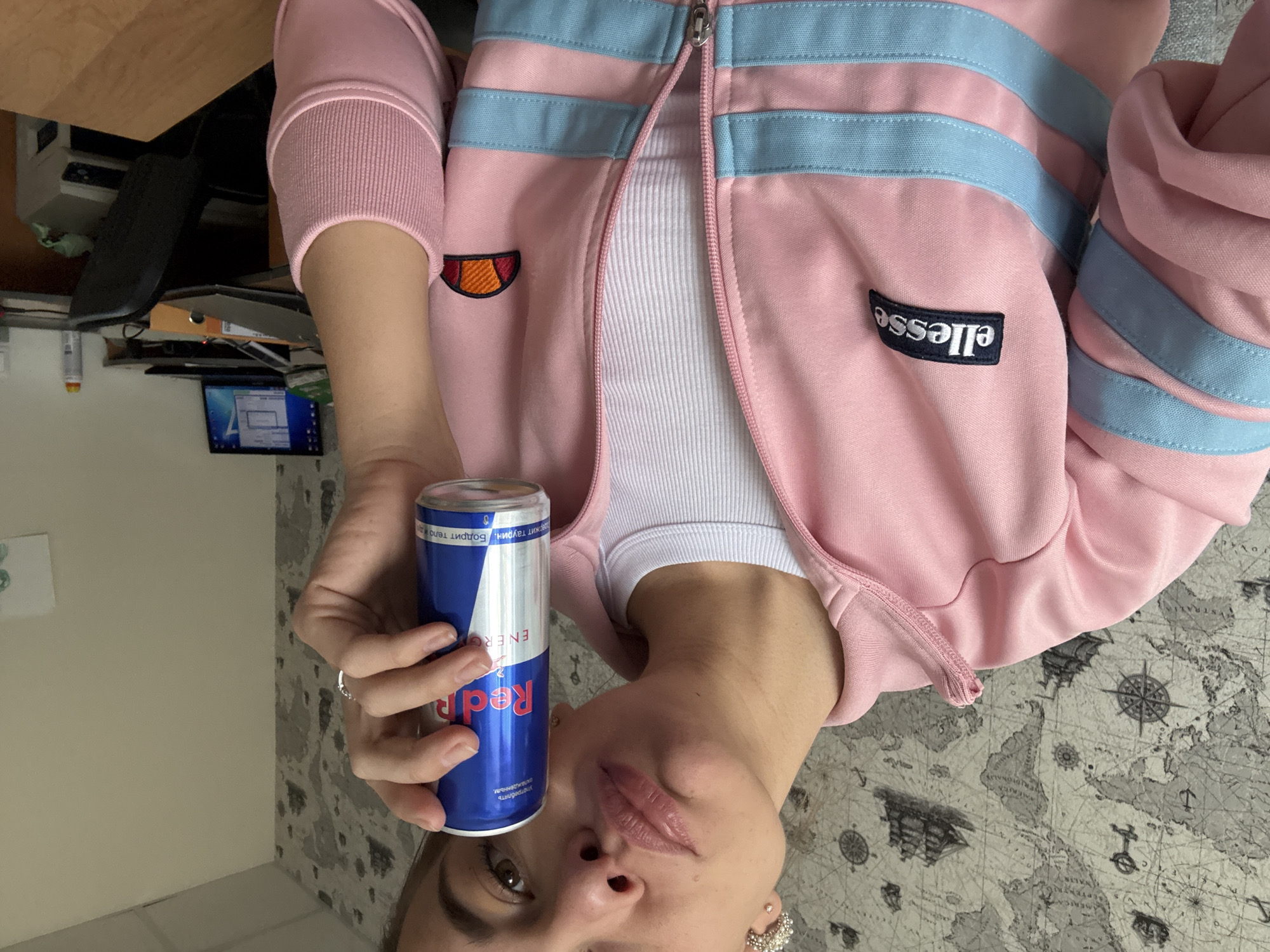 A person is pictured holding a blue and silver can of Red Bull energy drink. The person is wearing a pink track jacket with light blue stripes and a white tank top underneath. The background consists of a desk with a computer monitor, a black object, and some papers. The surface below is a gray patterned rug. This image does not contain enough information to determine a location in the city of Ust-Ilimsk, Russia.